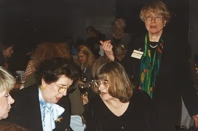 Drei Frauen in formeller Kleidung im Gespräch bei einer gesellschaftlichen Veranstaltung in einem dunklen Raum.