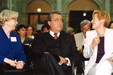 Drei Personen in formeller Kleidung sitzen in Stuhlreihen und unterhalten sich in einem Veranstaltungsraum.