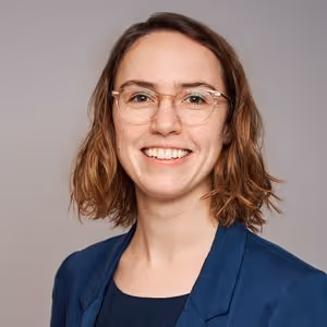 Porträt einer lächelnden Frau mit kurzen braunen Haaren, Brille und blauem Blazer vor grauem Hintergrund.