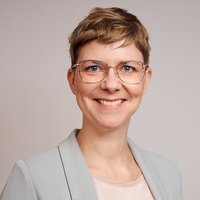 Porträt einer lächelnden Frau mit kurzen blonden Haaren und Brille, die einen hellgrauen Blazer trägt.