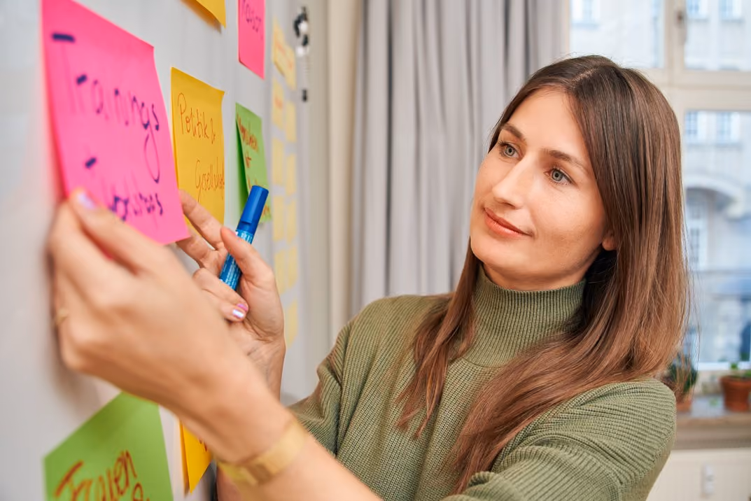 Frau klebt bunte Haftnotizen an eine Wand im Büro und hält einen blauen Stift.