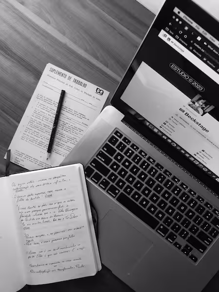 Overhead black and white photo of a laptop showing a website, an open notebook with handwritten notes, and a document titled 'SUPLEMENTO DE TRABALHO' with a pen resting on it, all placed on a wooden surface.