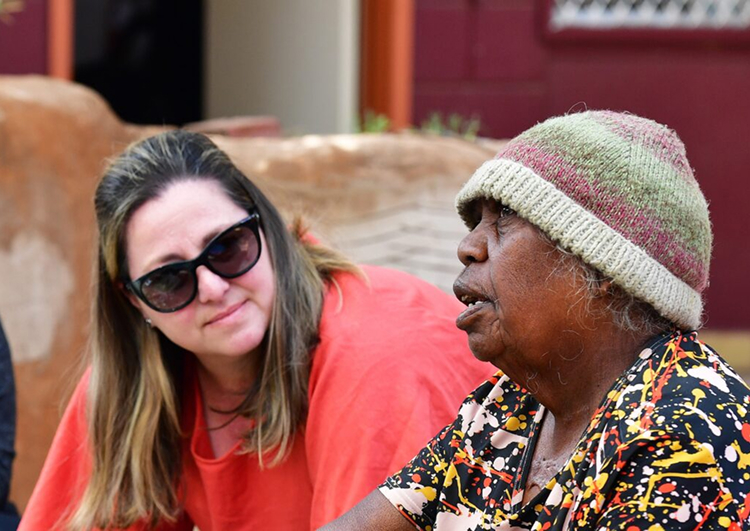 A woman in sunglasses and a red shirt listens attentively to an elderly woman wearing a knit hat and patterned shirt.