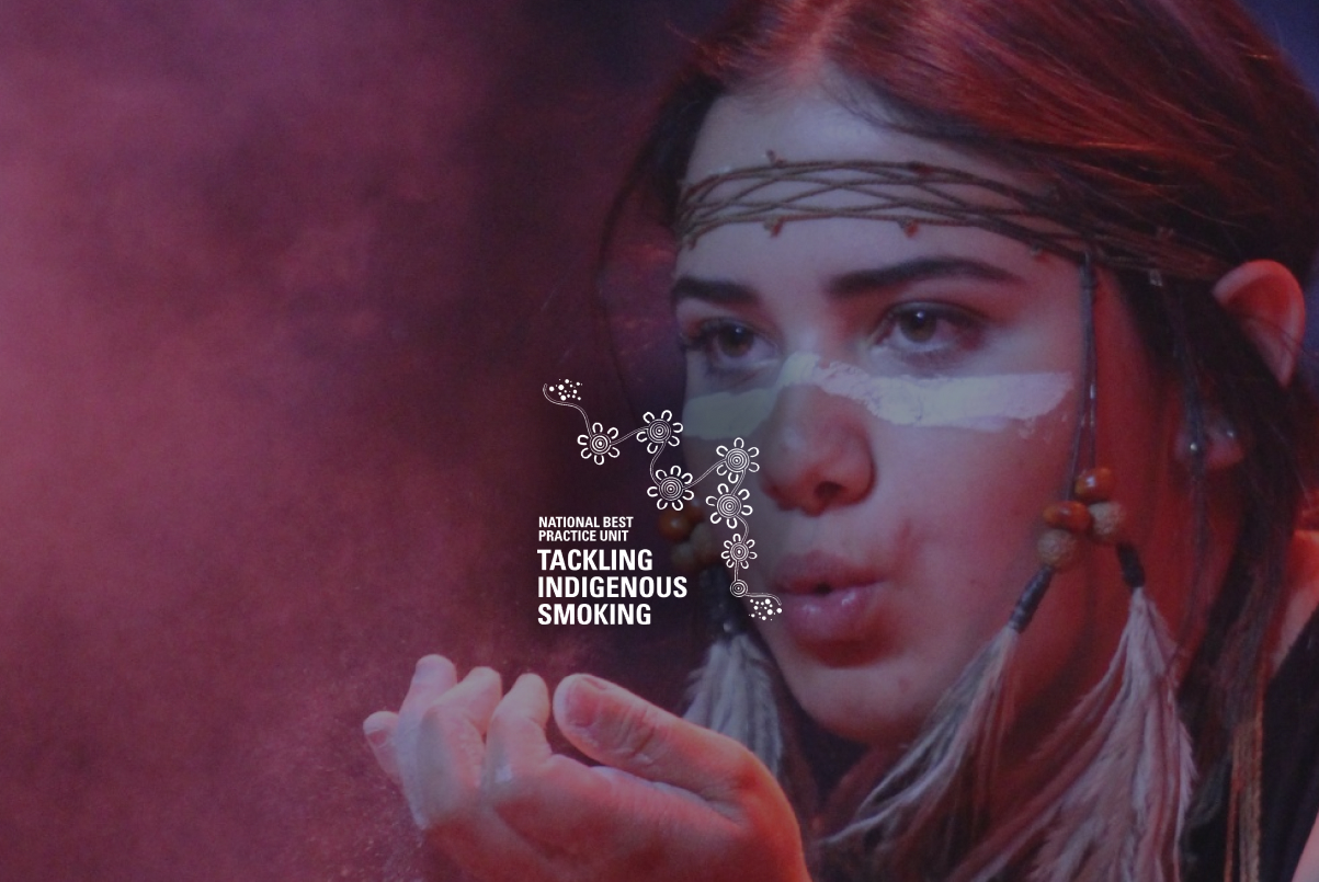 Young Indigenous woman with tribal face paint and braided hair blowing powder from her hands against a dark background.