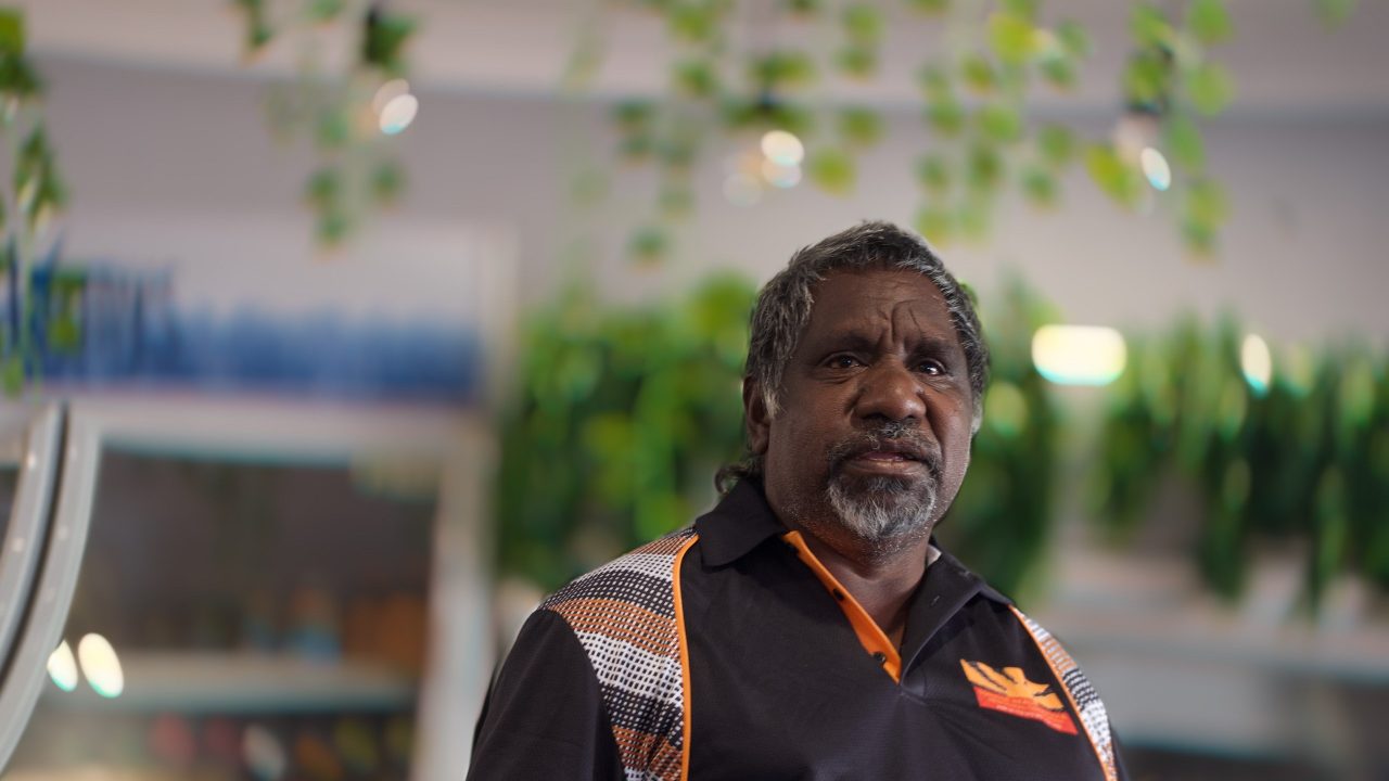 Portrait of an older man with salt-and-pepper hair and beard wearing a black and orange patterned shirt indoors with blurred green plants in the background.