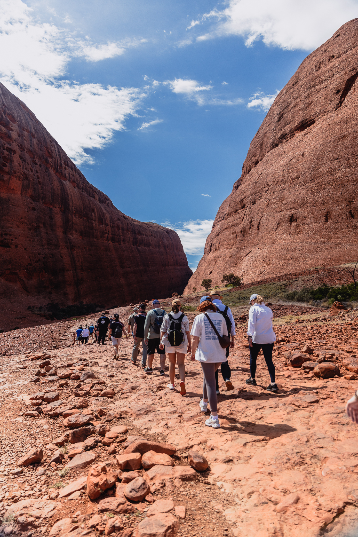 Group of hikers walking on a rocky, red desert trail between two large red rock formations under a blue sky with some clouds.