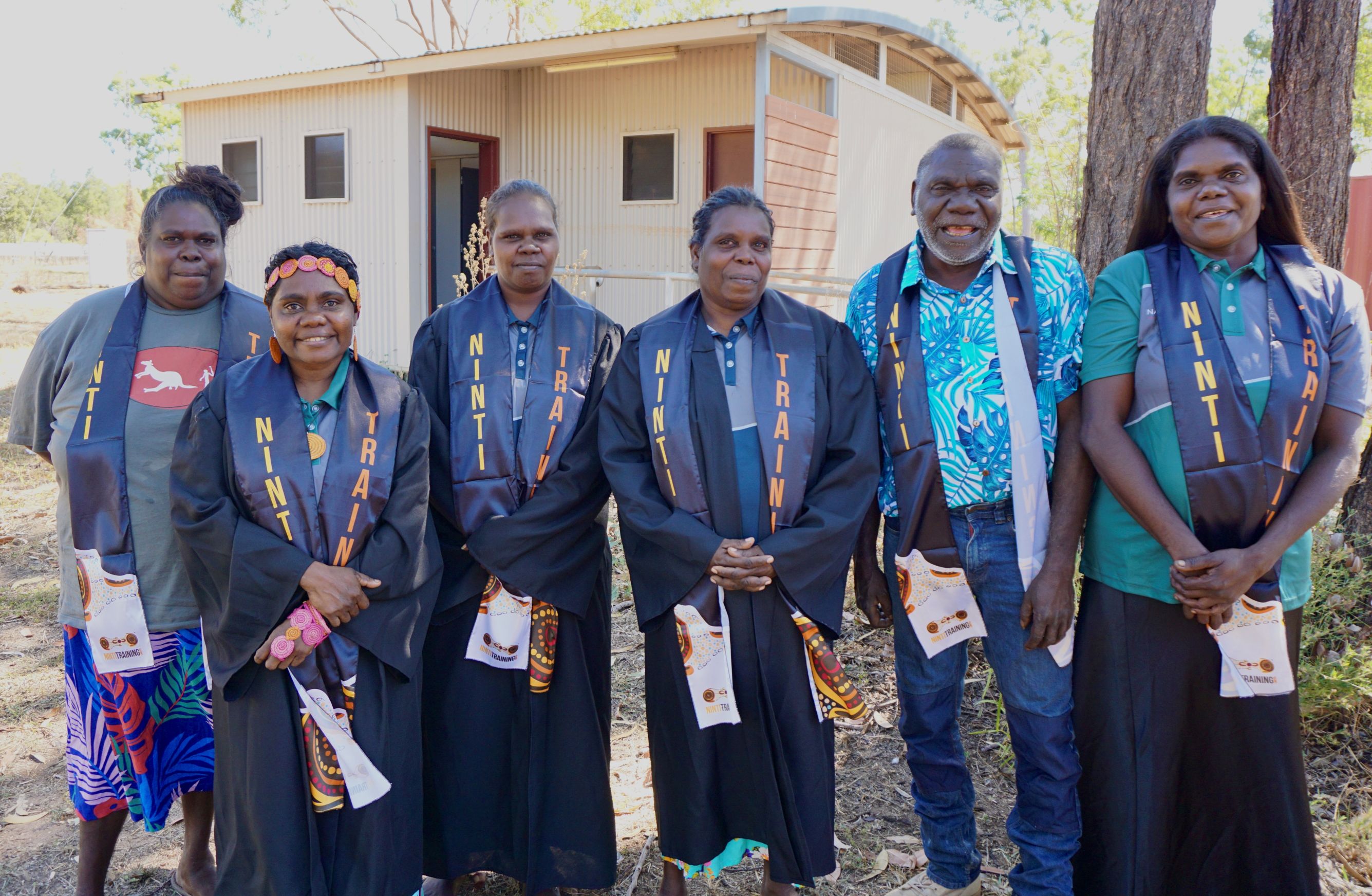 Group of six Indigenous adults standing outside a building wearing black gowns and stoles labeled 'Ninti Training', smiling at the camera.
