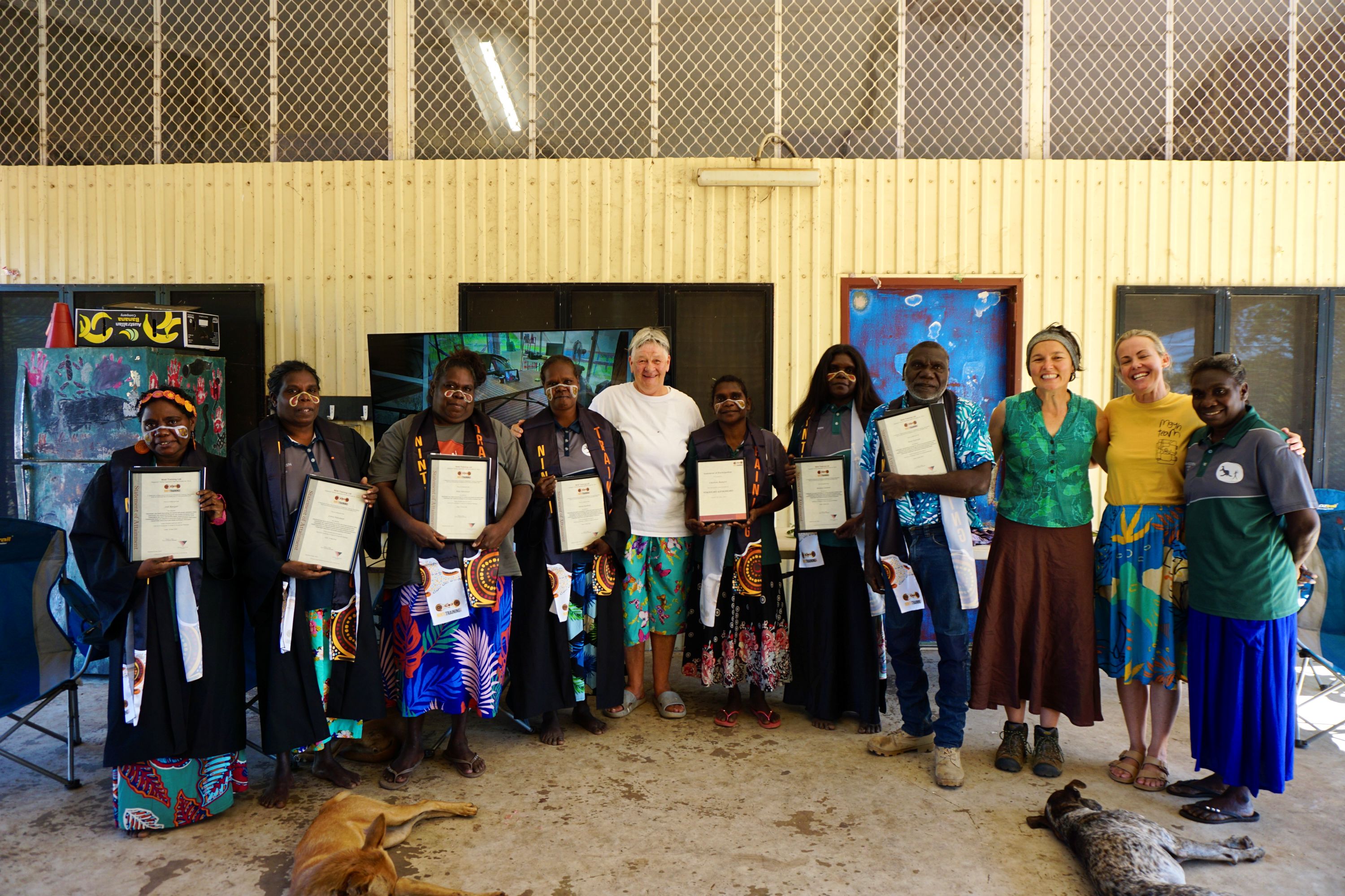 Group of Indigenous people and two women standing indoors, holding certificates, with two dogs lying on the floor.