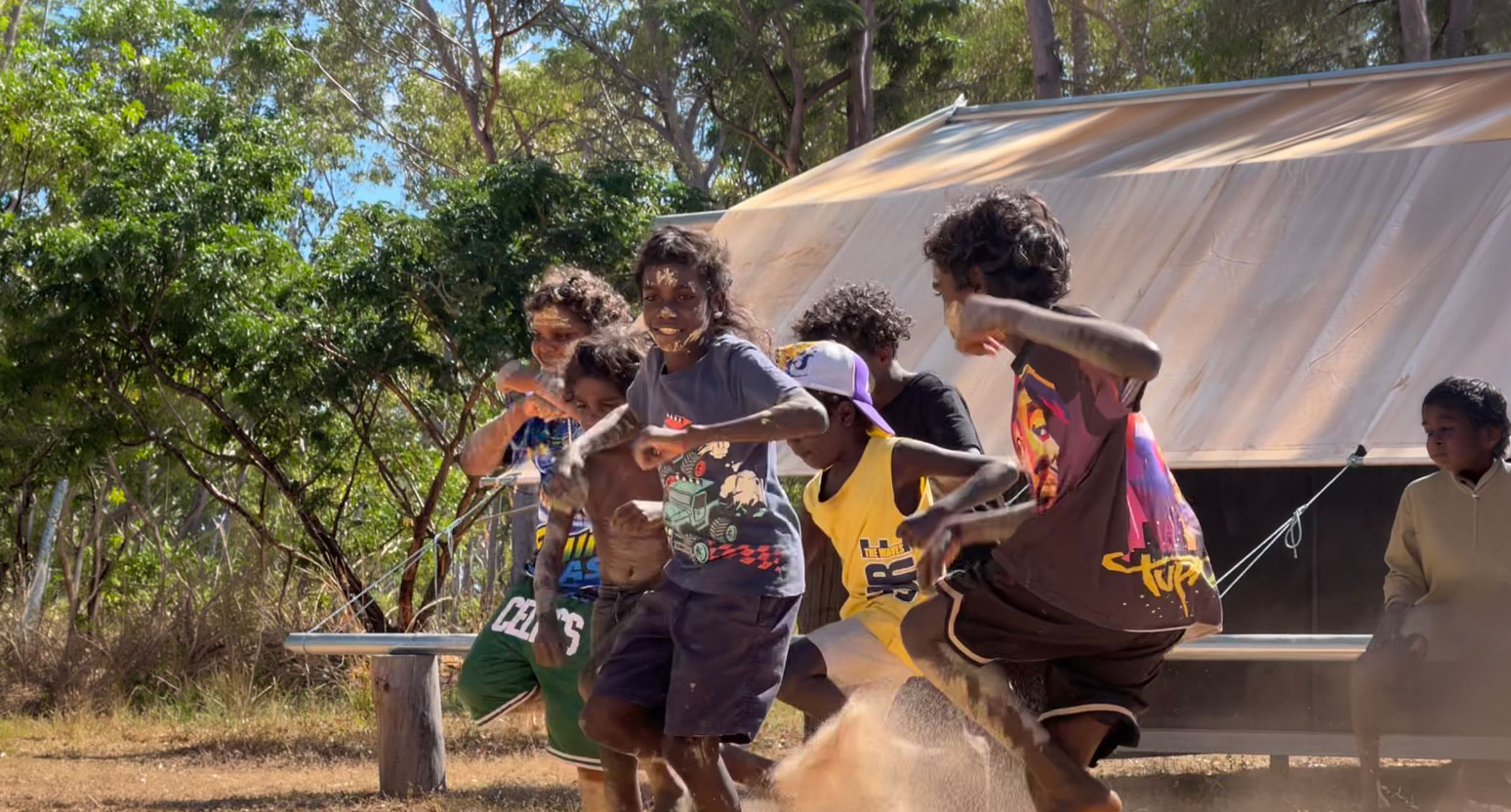 Group of children with painted faces playing energetically outdoors near a tent and trees.