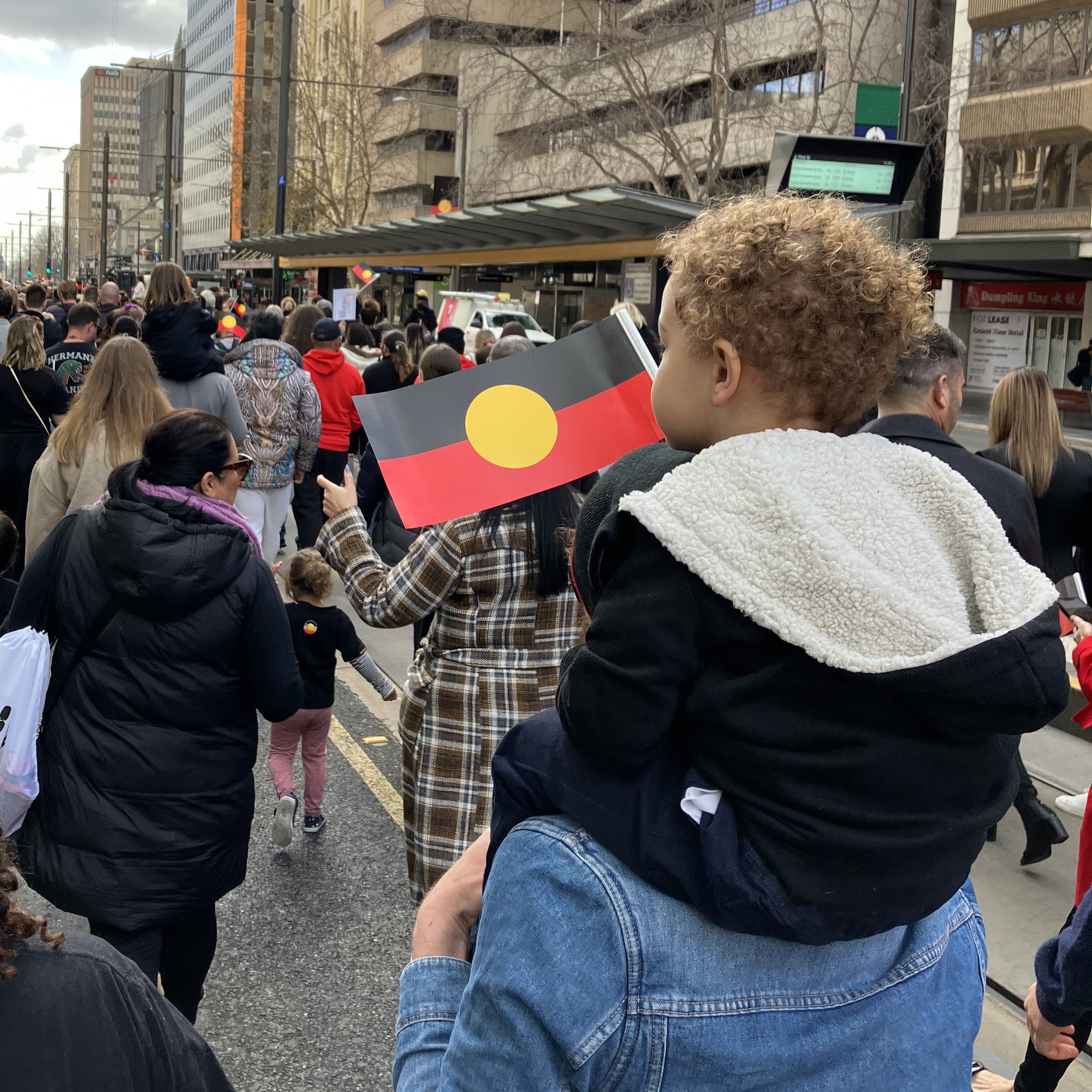 Crowd of people walking on a city street during a march, a child on an adult's shoulders holding an Aboriginal flag.