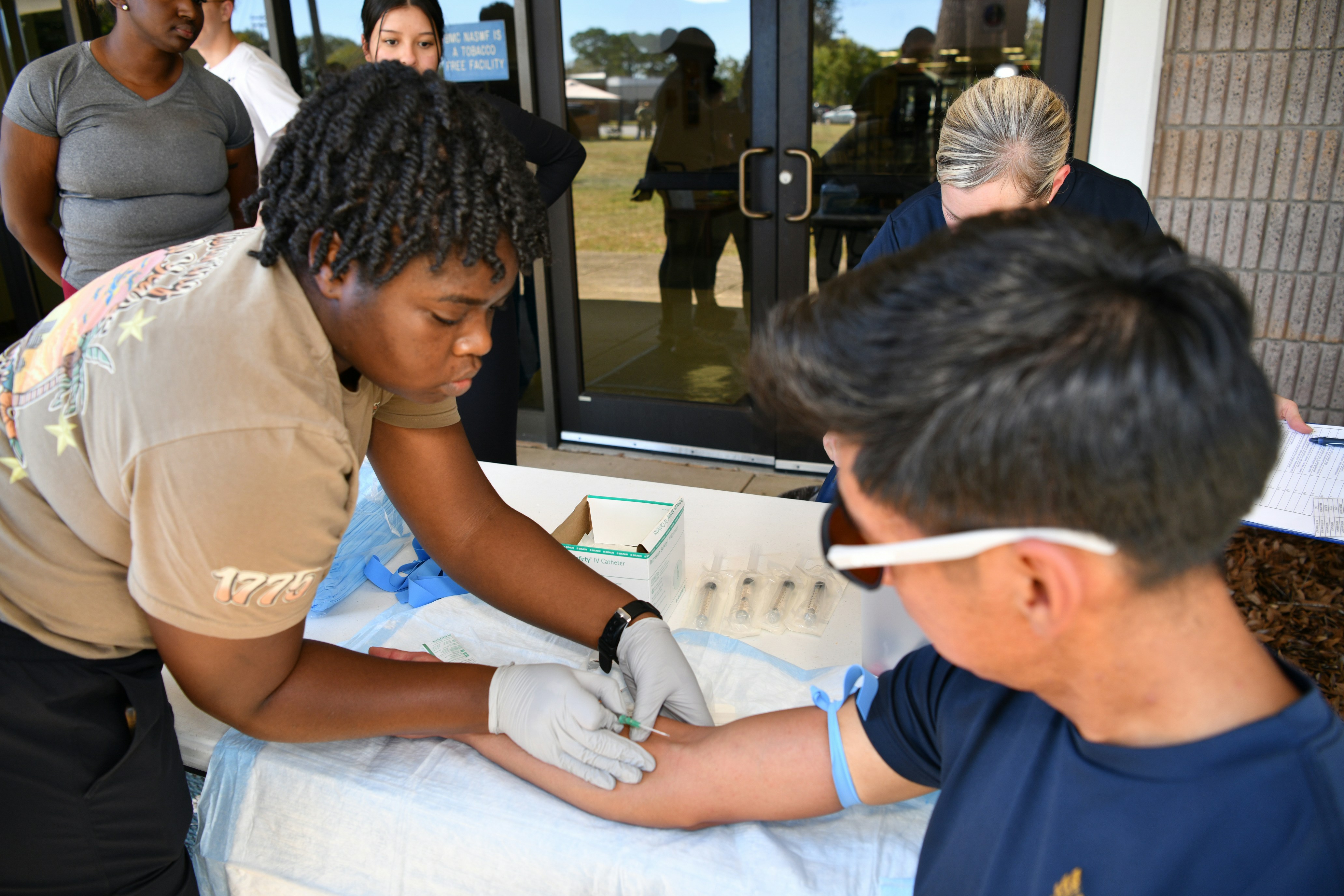 Healthcare worker wearing gloves inserting a needle into a person's arm at a medical testing or vaccination site outdoors.