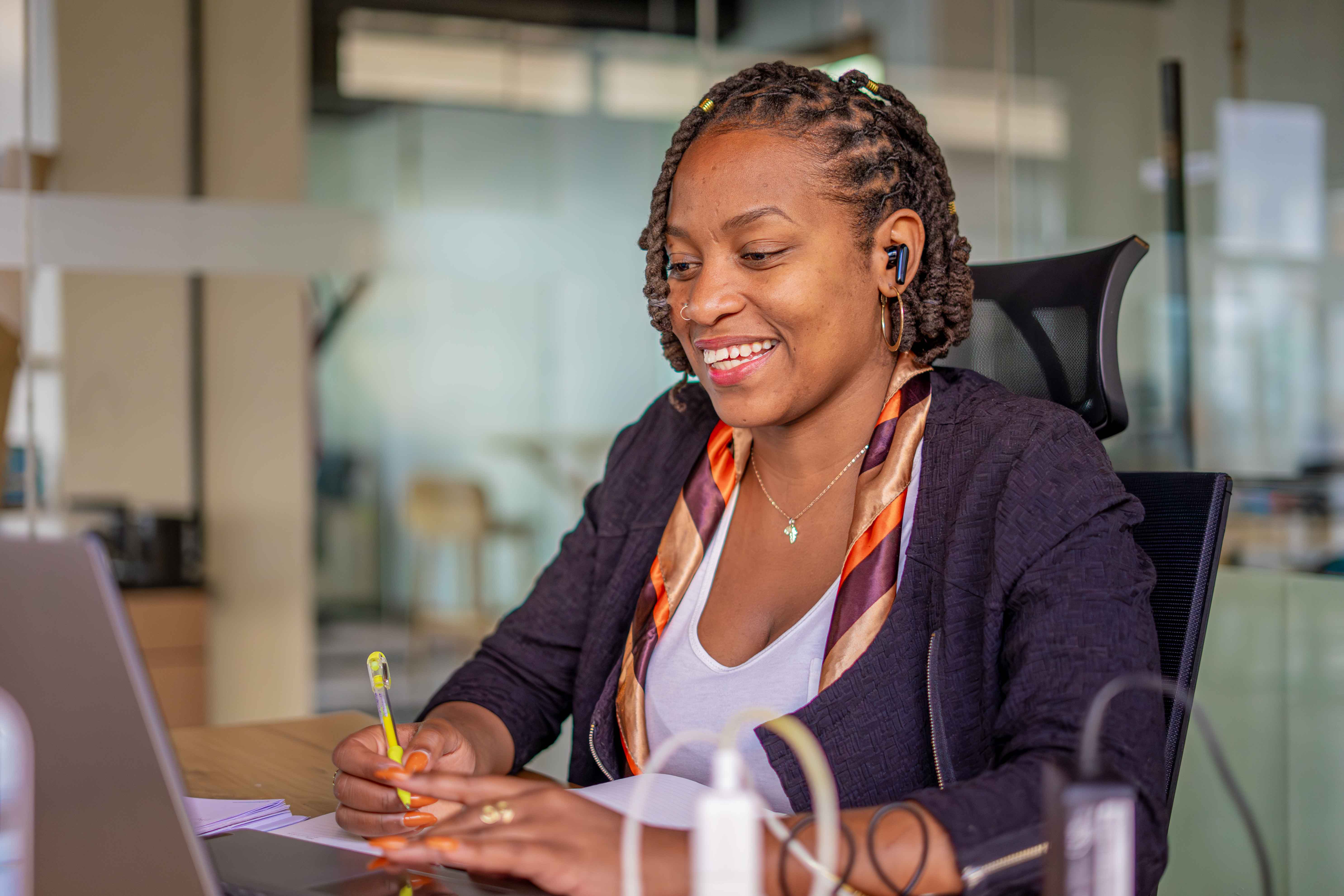 Natalie Mukami; Smiling woman with braided hair and earbuds working on a laptop at a desk in an office.