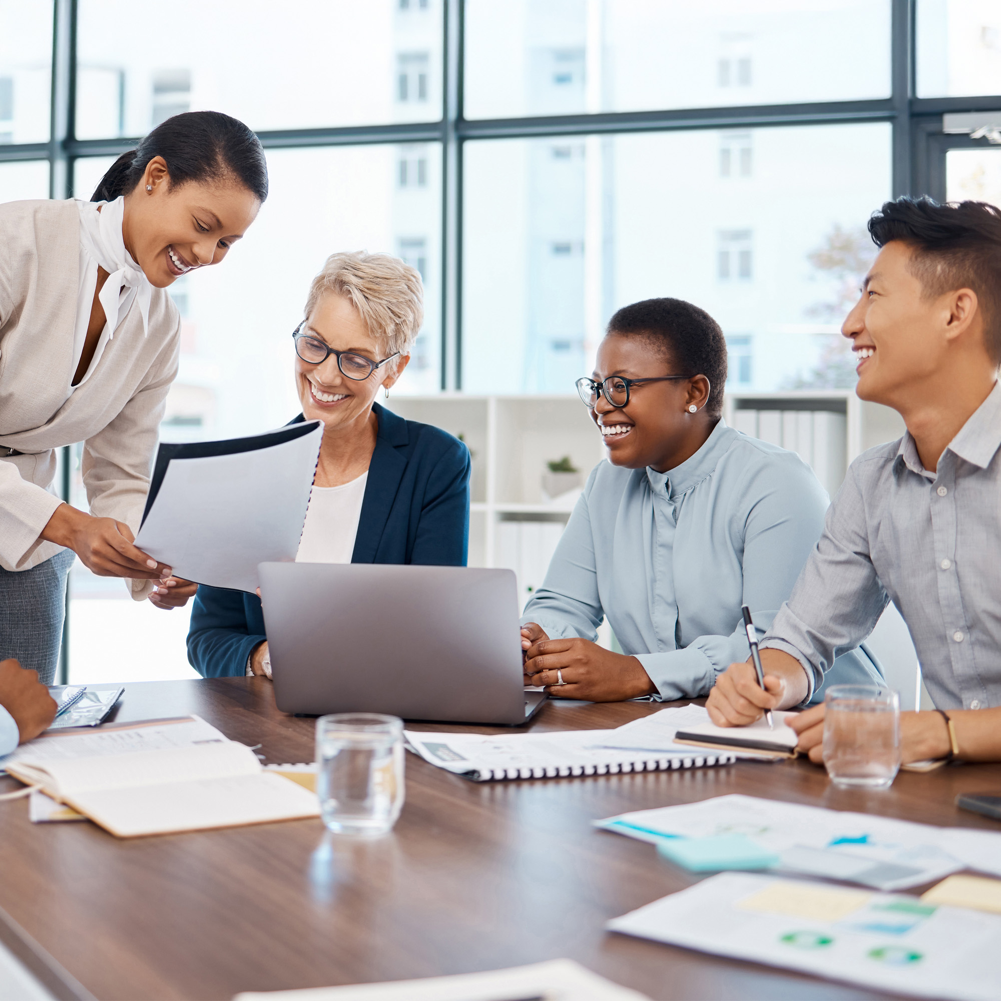 Four diverse professionals laughing and discussing documents during a meeting around a conference table with a laptop and papers.