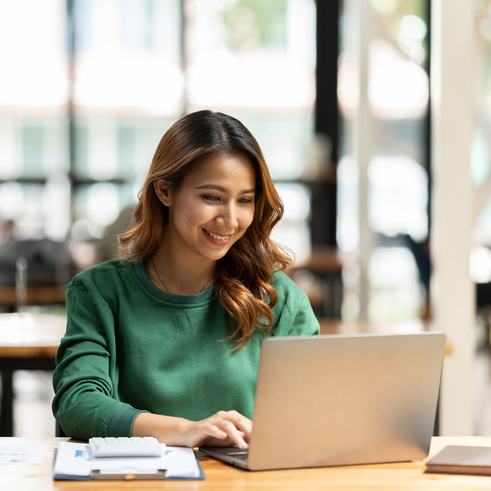 Young woman in a green sweater smiling while working on a laptop at a wooden desk in a bright office.