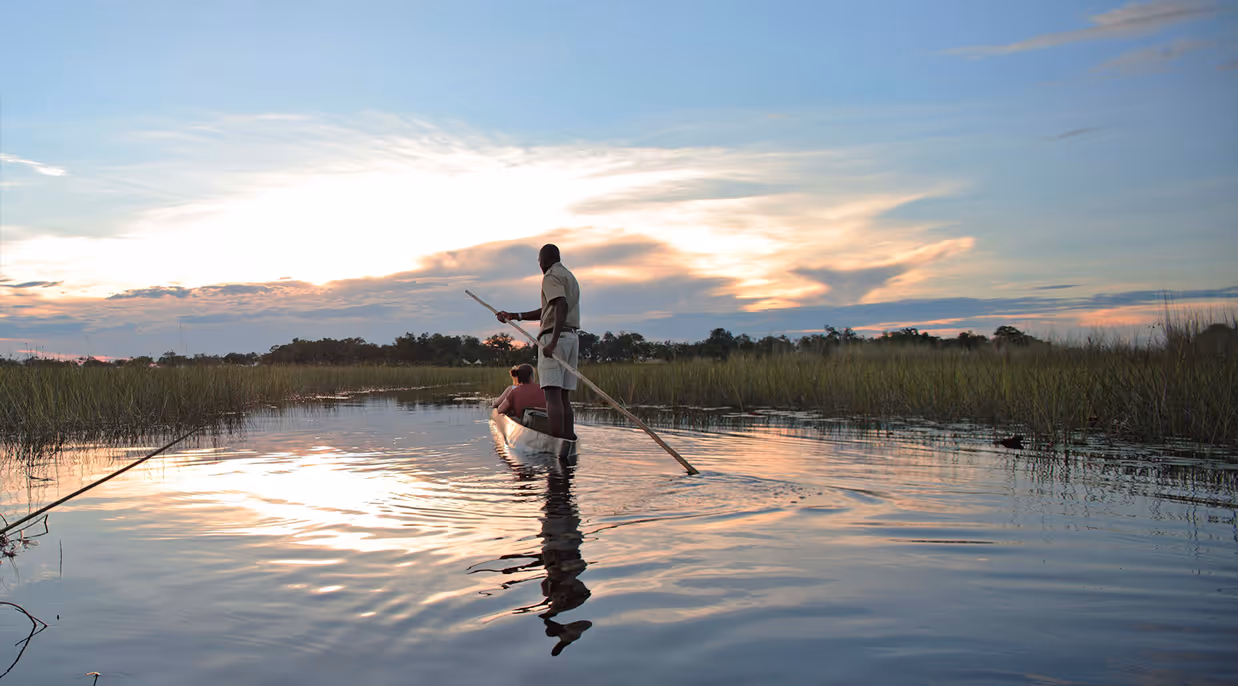 Two people in a canoe on calm water at sunset with tall grasses on both sides.