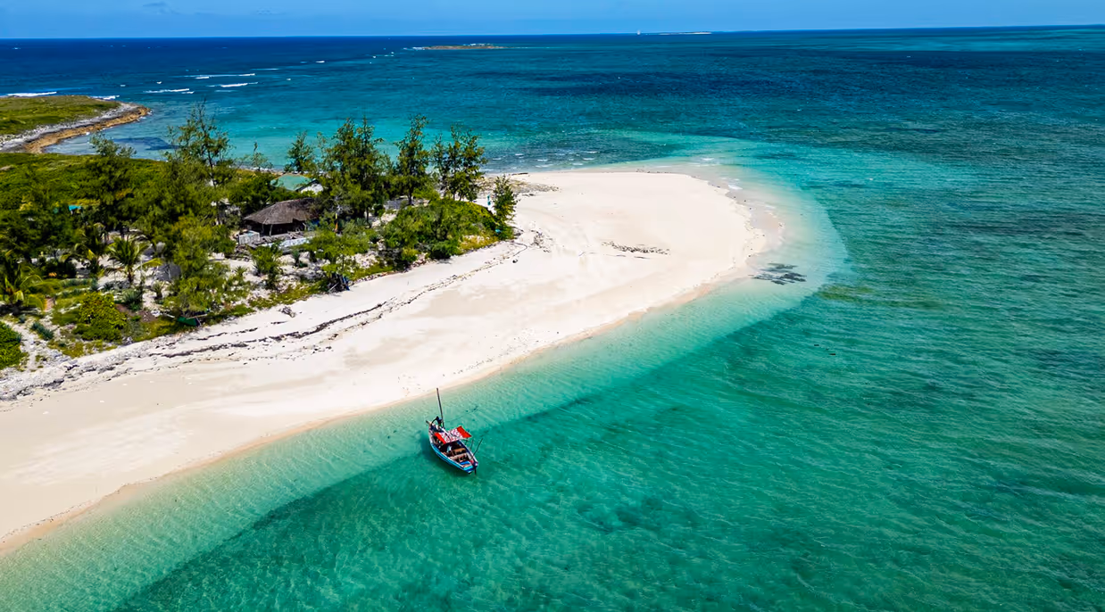 A small boat floats near a white sandy beach with green trees and turquoise ocean water.