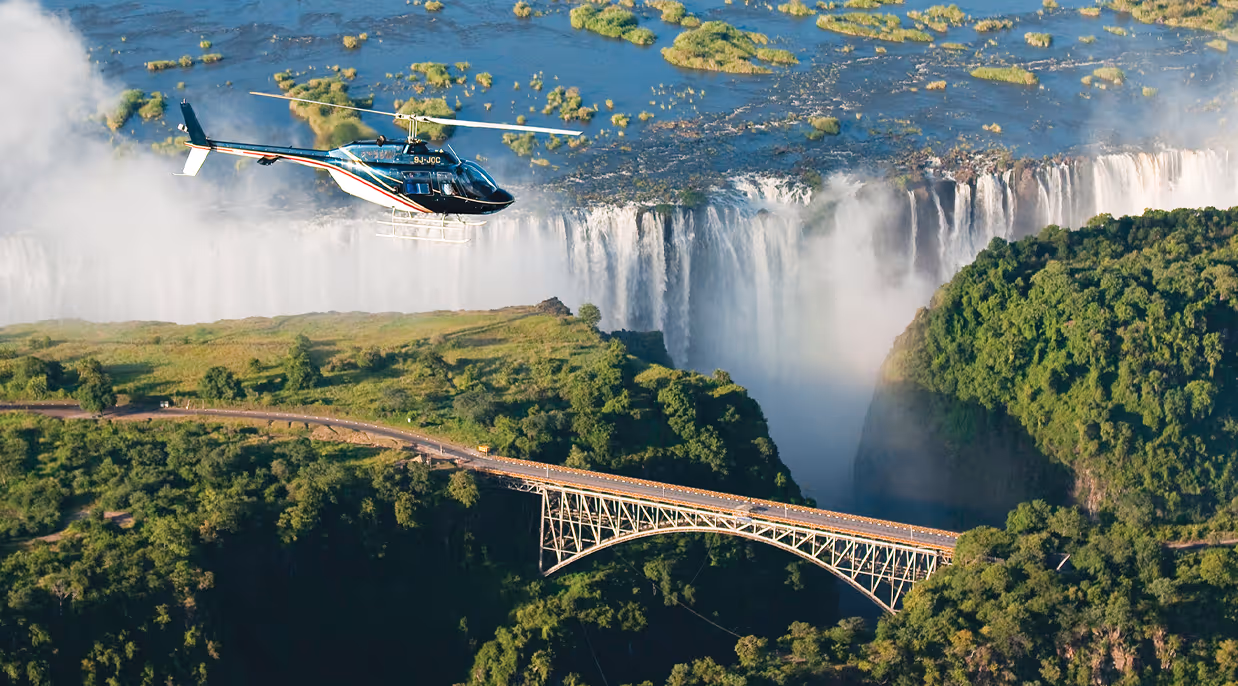 Helicopter flying near a lush green landscape with a bridge and large waterfalls in the background.
