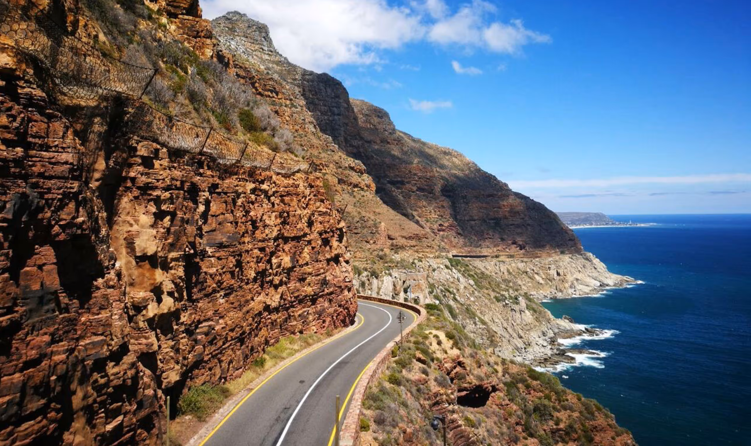 Winding coastal road beside cliffs with blue ocean under a partly cloudy sky.