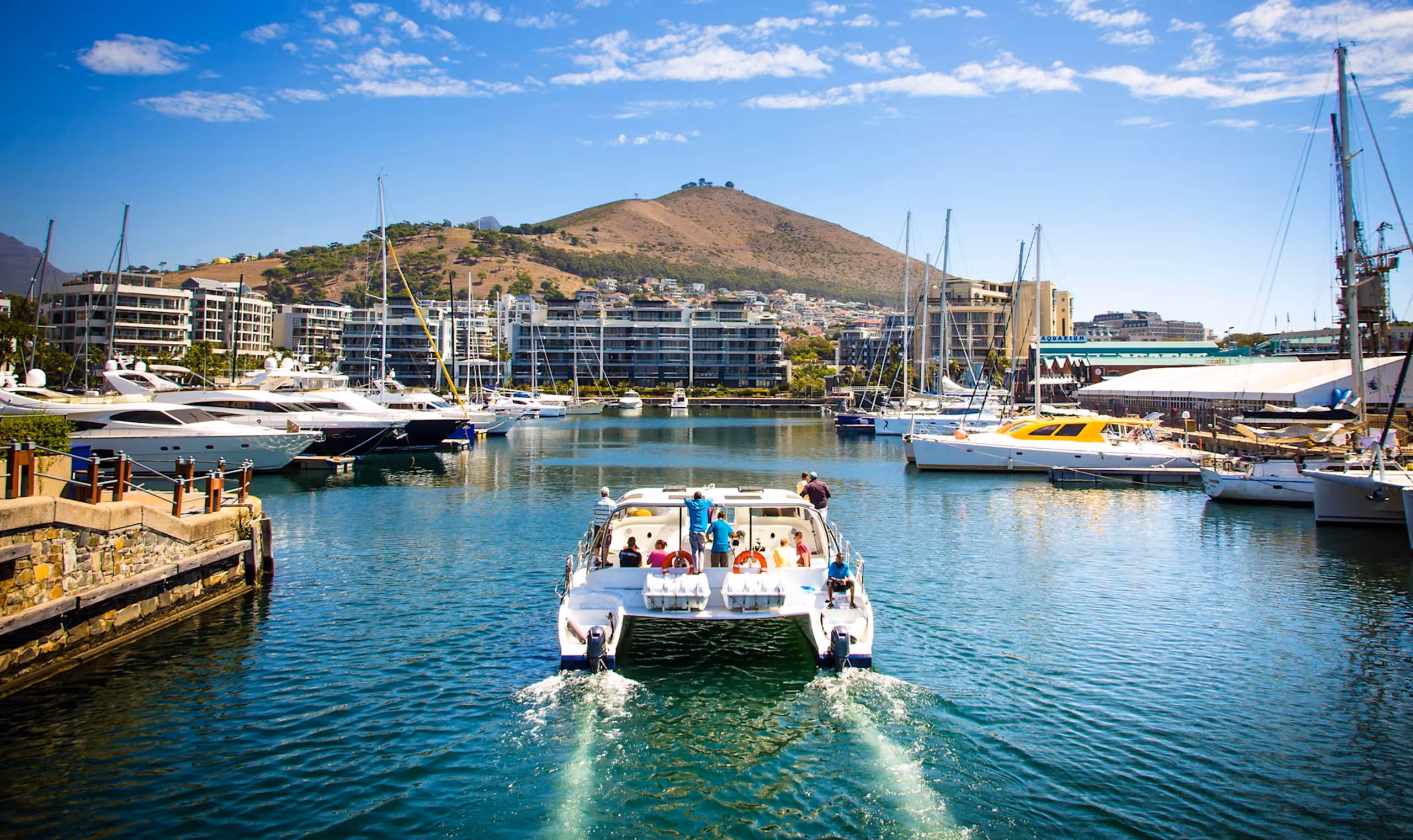 Boat with passengers cruising in a marina with yachts and hillside buildings under a blue sky.