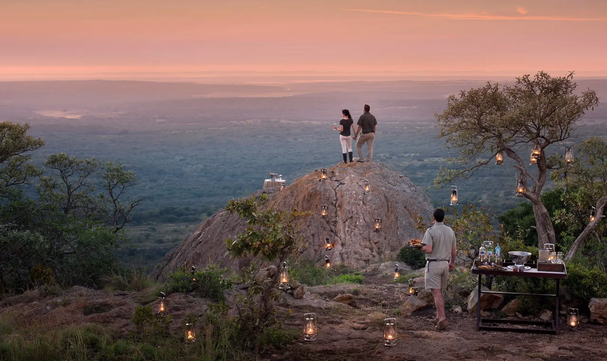 Couple holding hands on a rock overlooking a vast landscape at sunset with lanterns lit around.