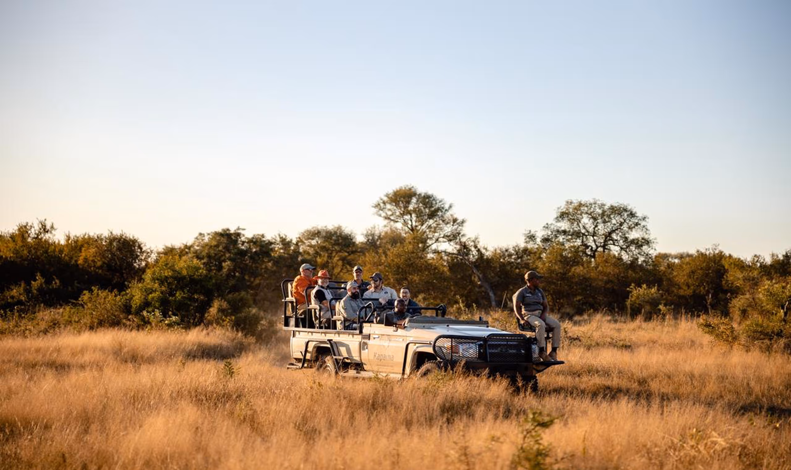 Group of people on open safari vehicle driving through tall grass in a golden savanna landscape.