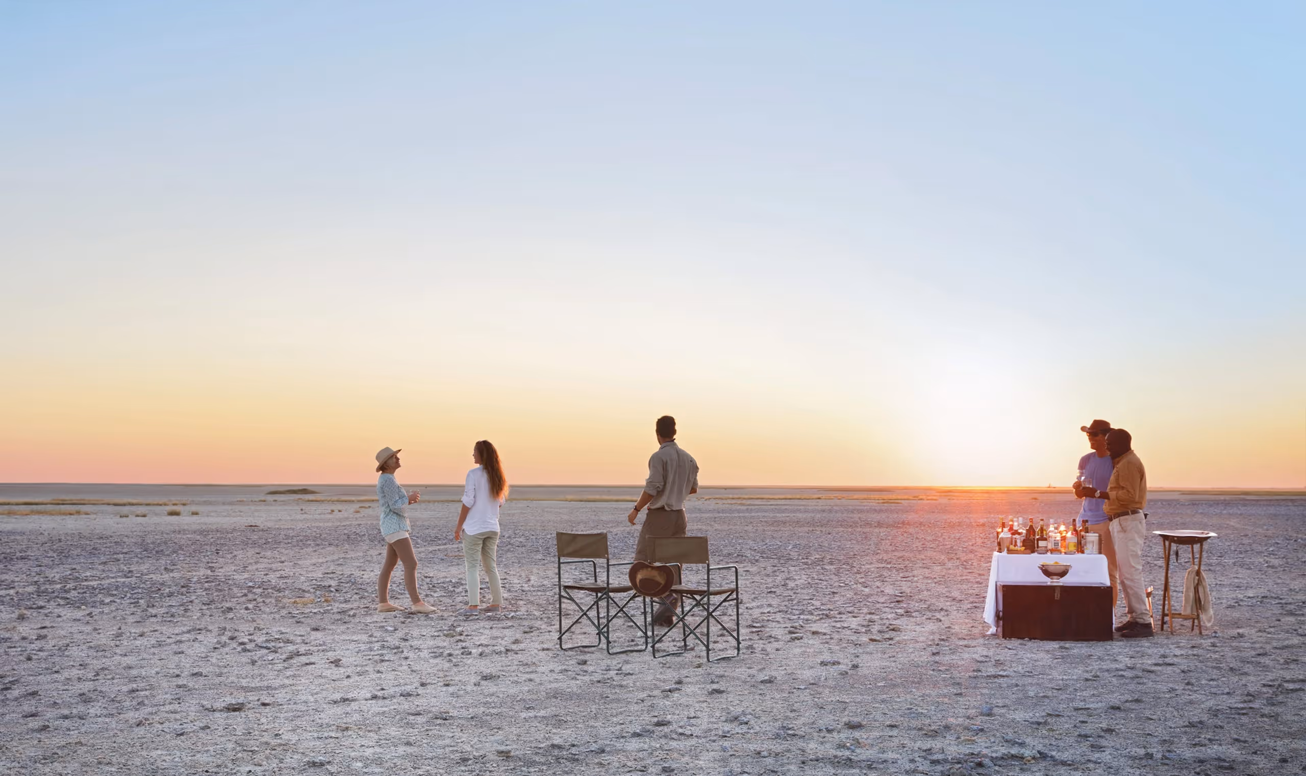 Five people standing and chatting near two chairs and a table with drinks on a salt flat at sunset.