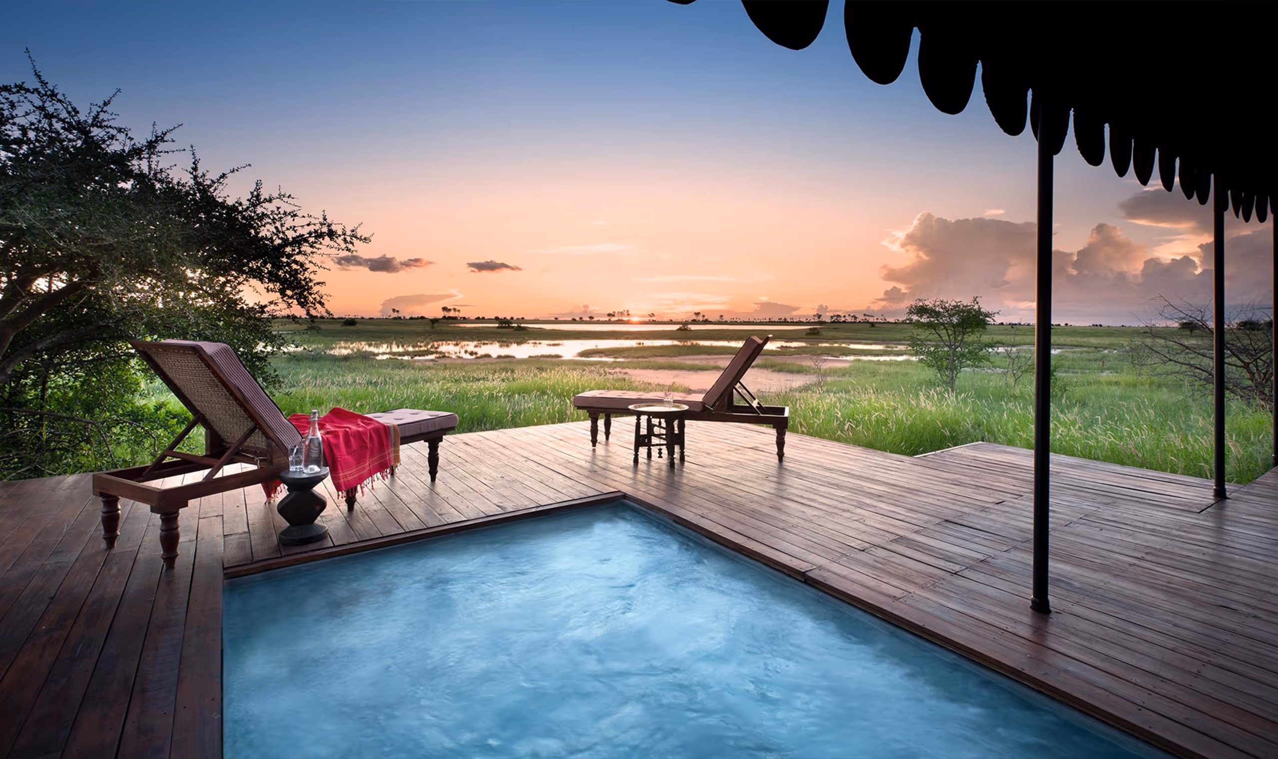 Wooden deck with pool, lounge chairs, and table overlooking green field at sunset.