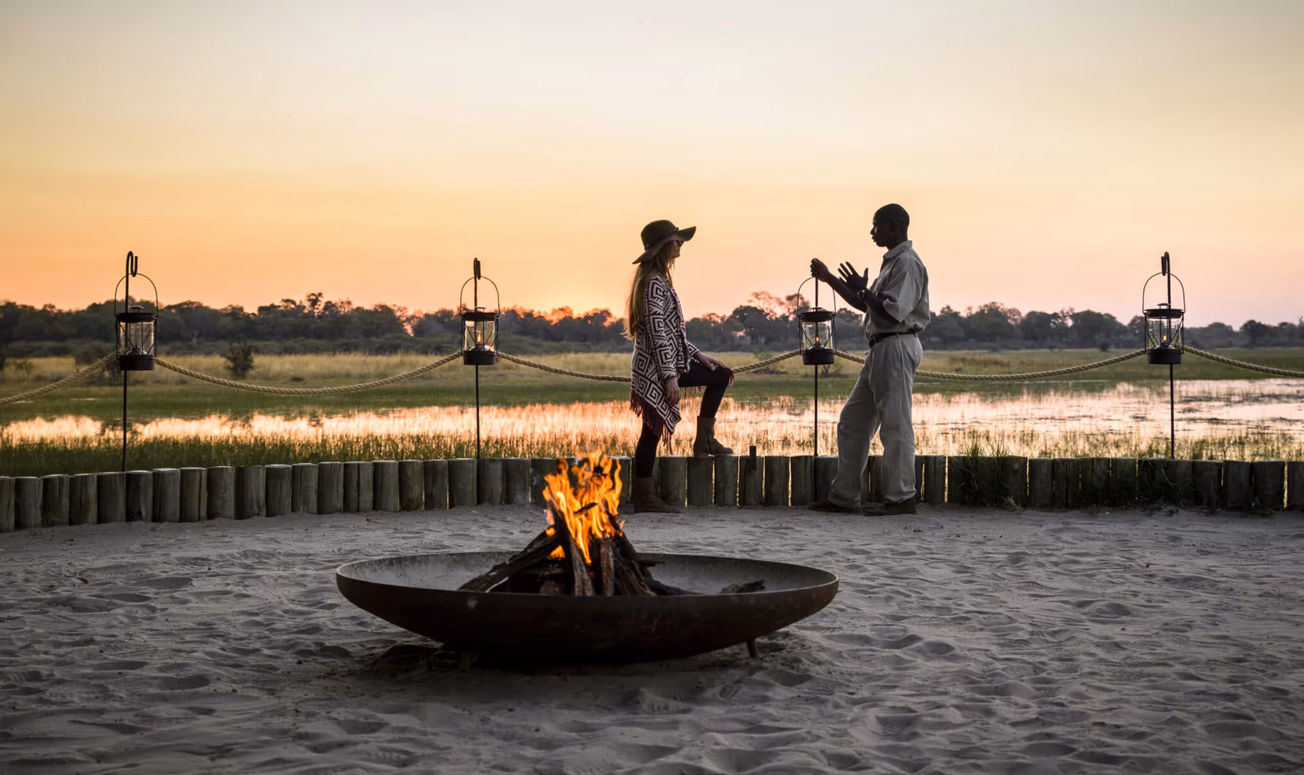 Two people talking by a campfire with lanterns near a water body at sunset in a safari setting.