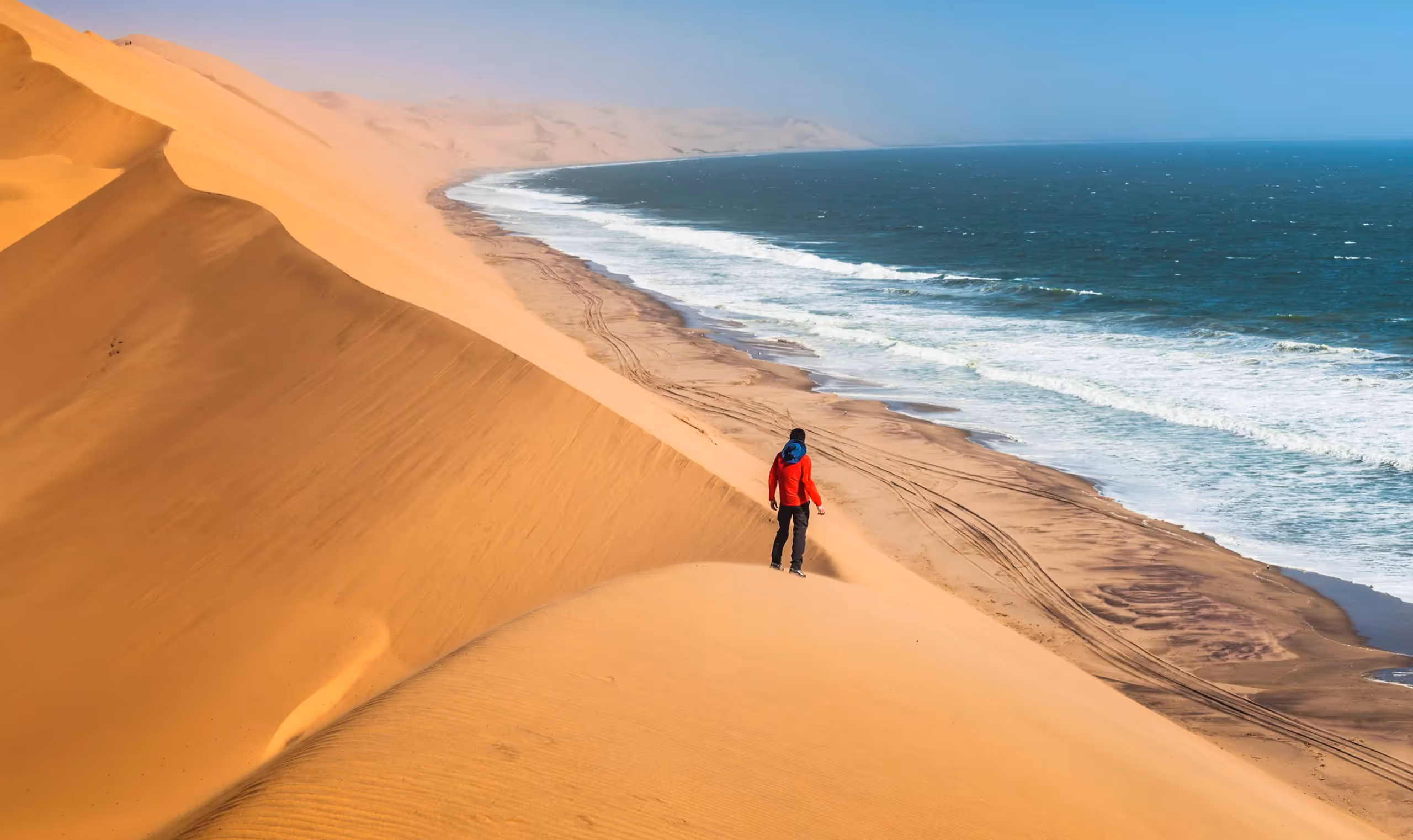 Person in red jacket standing on sand dunes near a beach with ocean waves under blue sky.