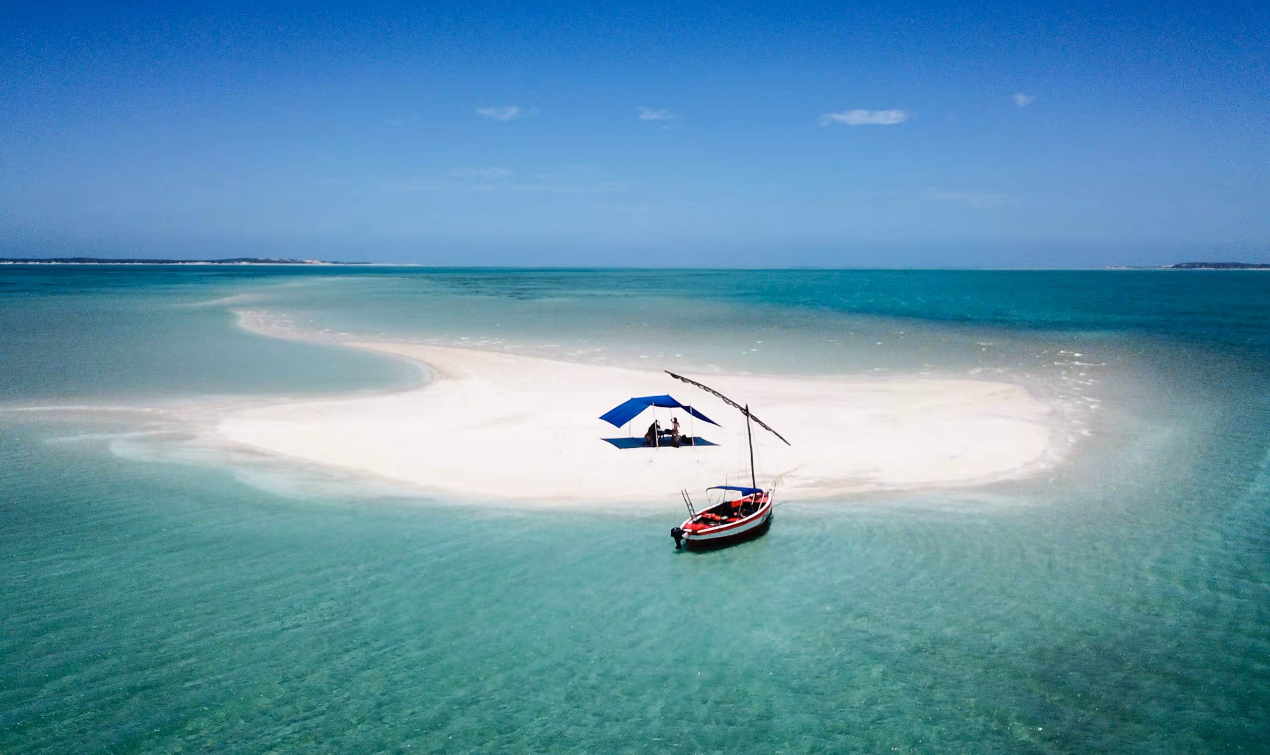 Small sandy island with a blue tent and red boat surrounded by clear turquoise ocean under blue sky.