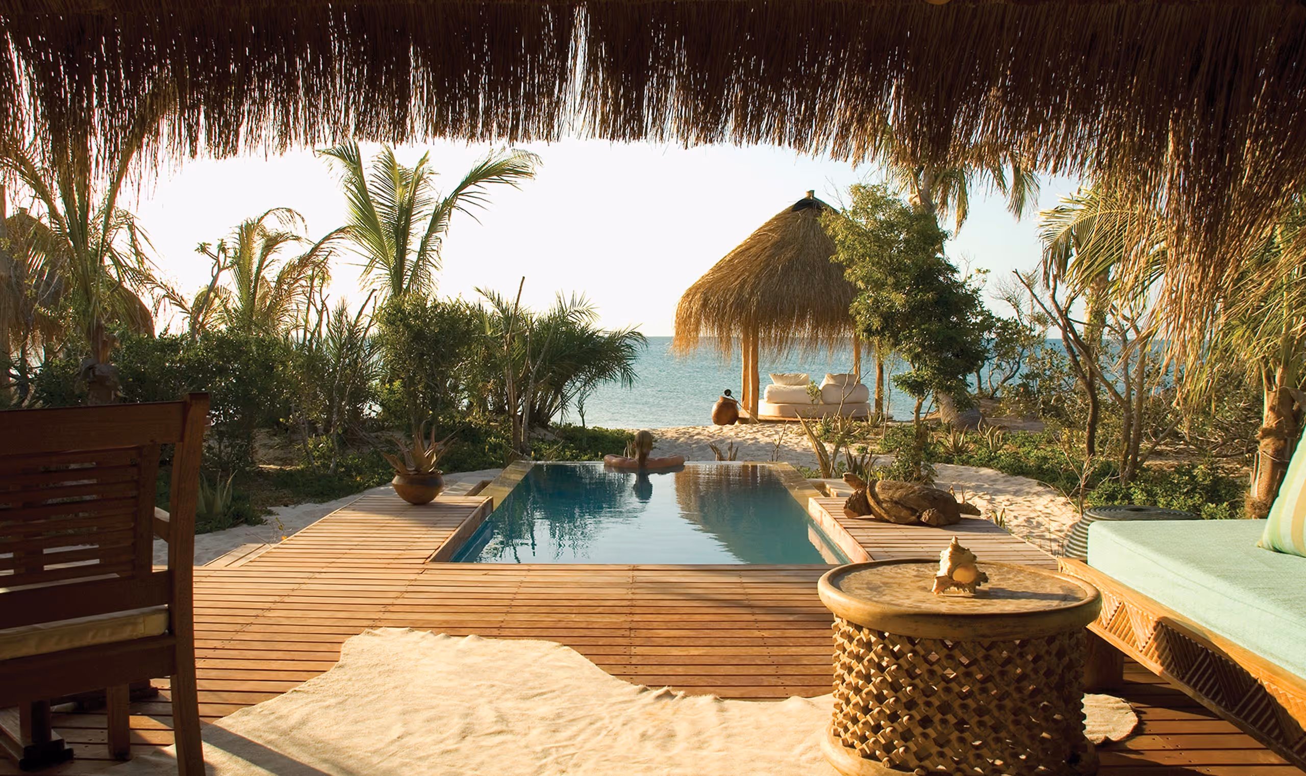 A person relaxing on a float in a pool overlooking a beach hut and ocean with tropical plants around.