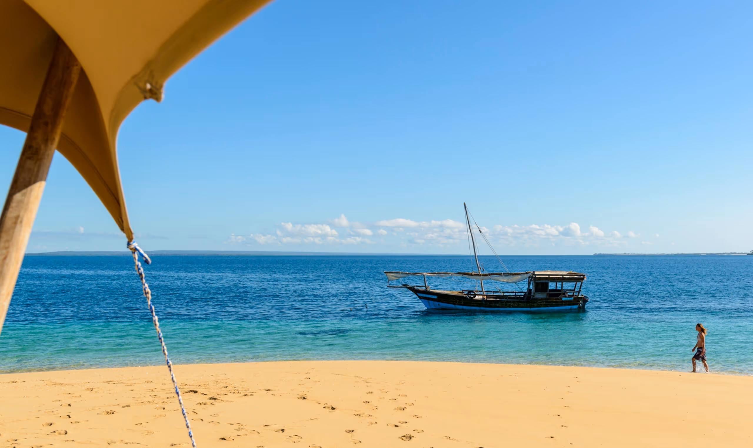 Sandy beach with a traditional wooden boat on clear blue water and a person walking at shore.