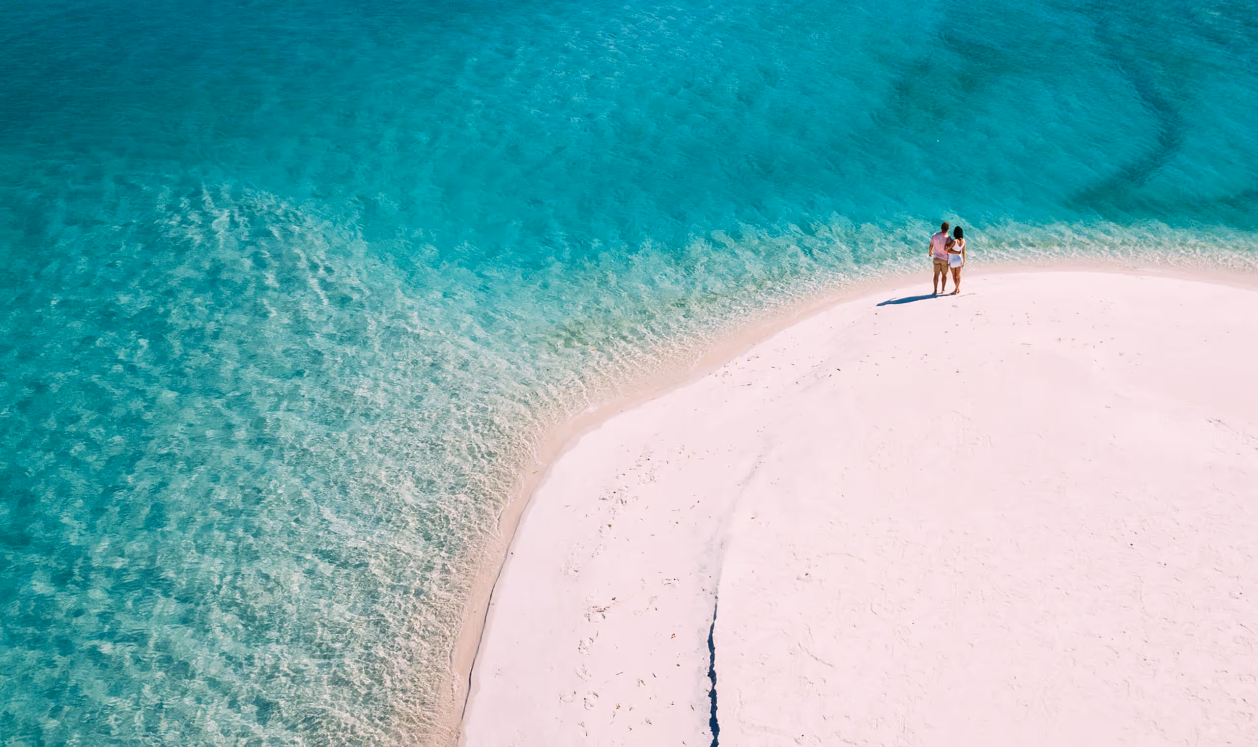 Couple standing on white sand beach next to clear turquoise ocean water.