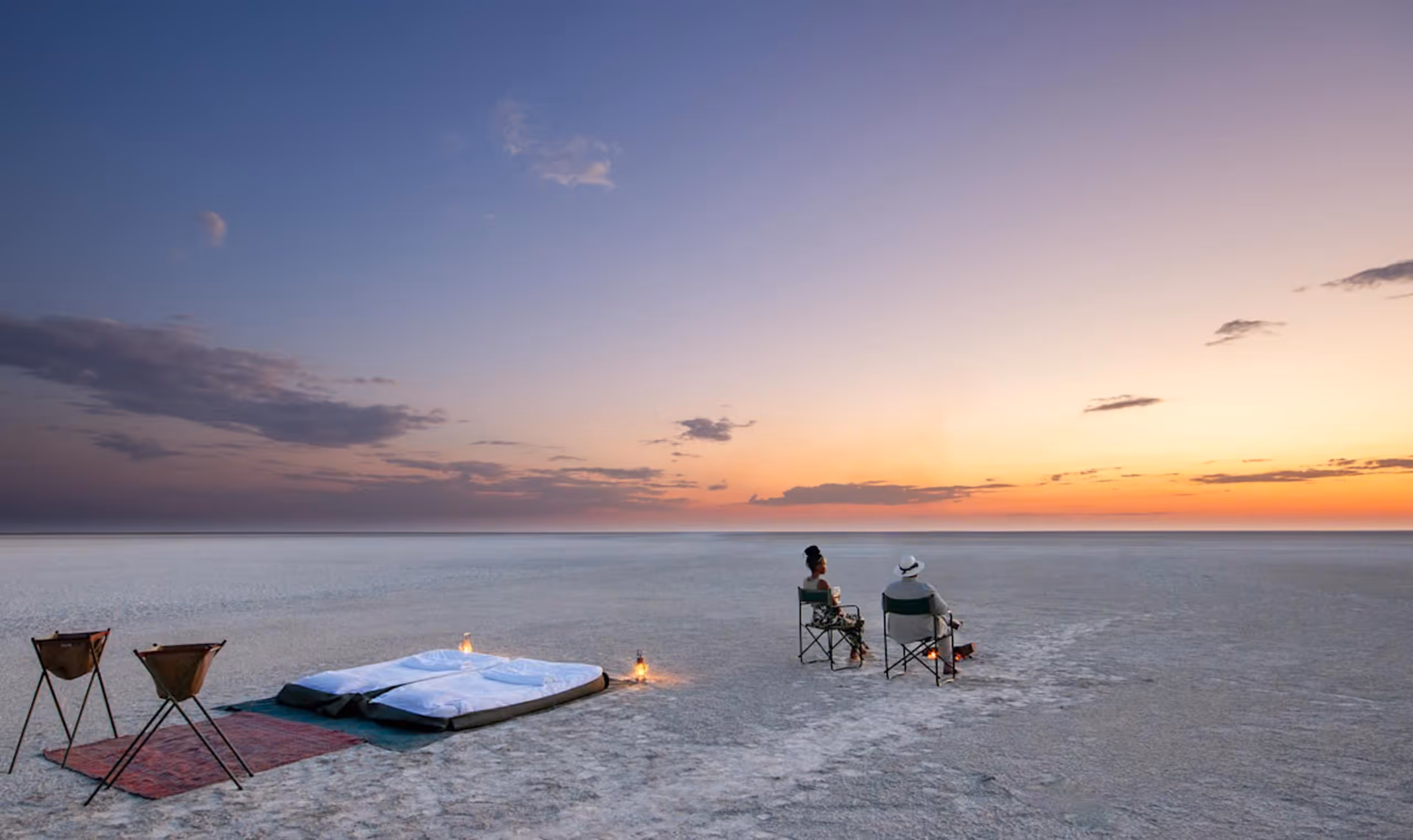 Two people sitting in chairs on flat land at sunset with bed and lanterns nearby.