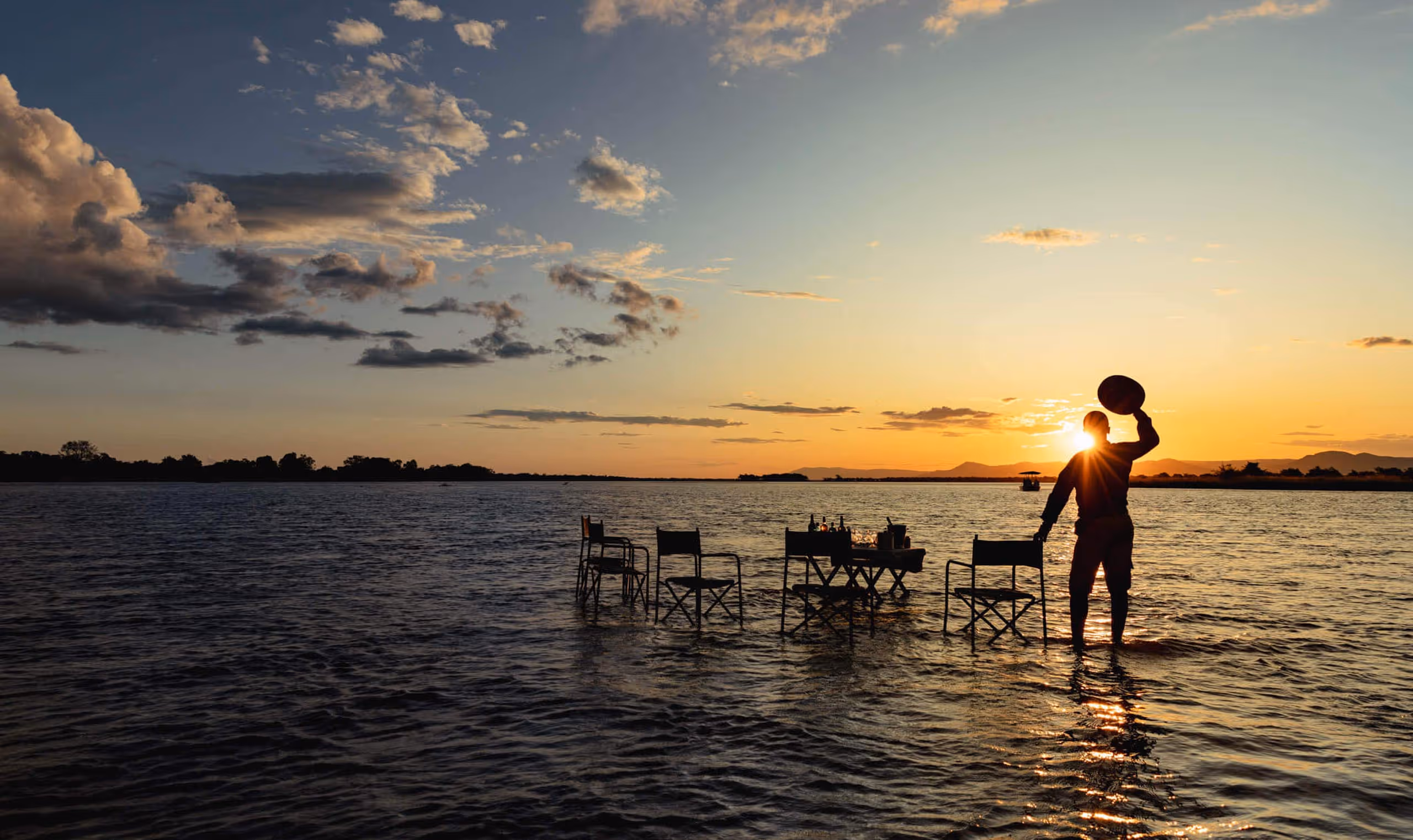 Silhouette of person holding hat near table and chairs set in shallow river at sunset.