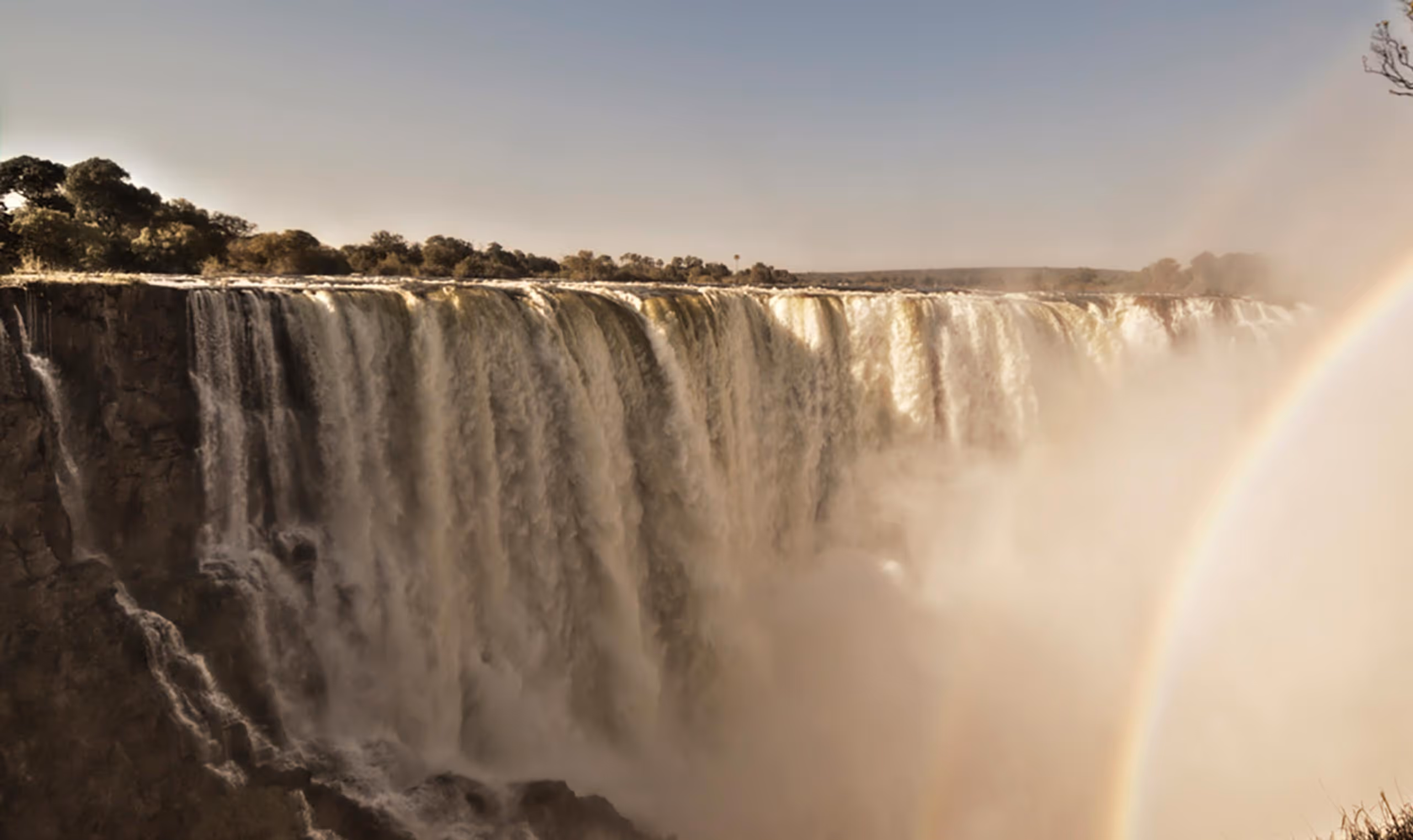 Wide view of Victoria Falls with water cascading and a faint rainbow in the mist.