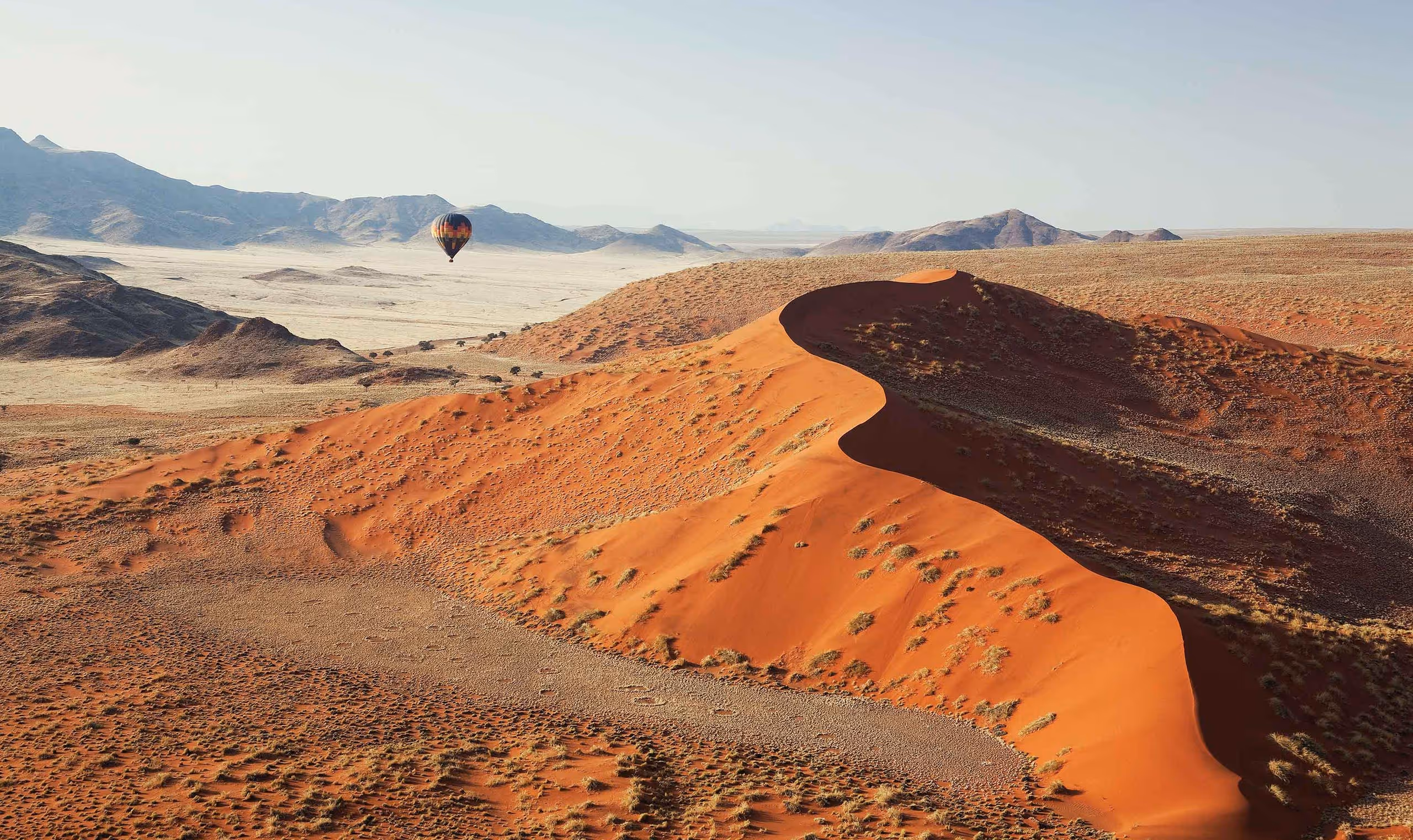 A colorful hot air balloon floats over orange sand dunes and desert landscape with mountains in the distance.