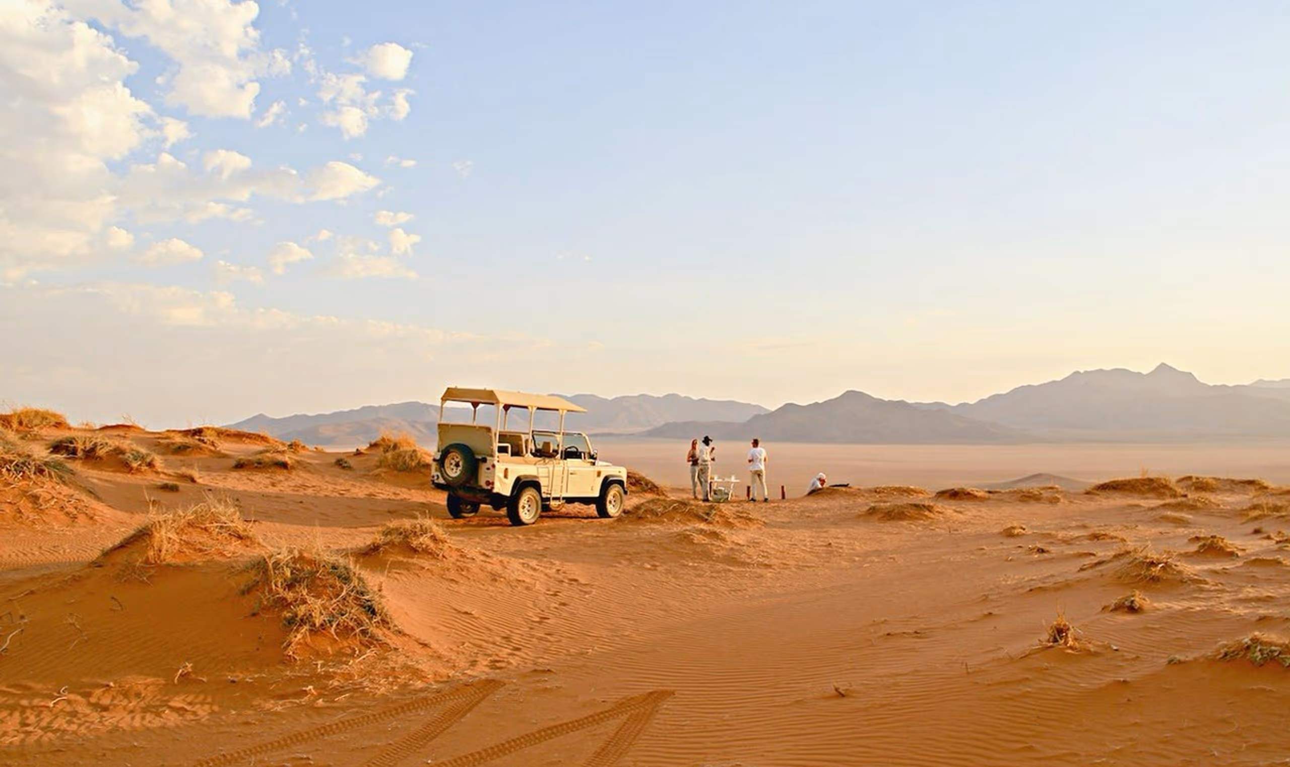 White safari jeep parked on sand dunes with people near table and mountains in background.