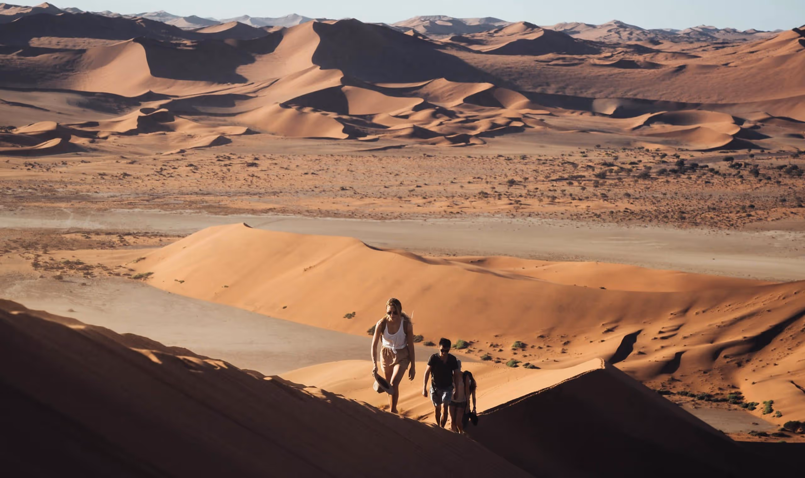 Three people hiking up large sand dunes in a vast desert landscape with distant mountains.