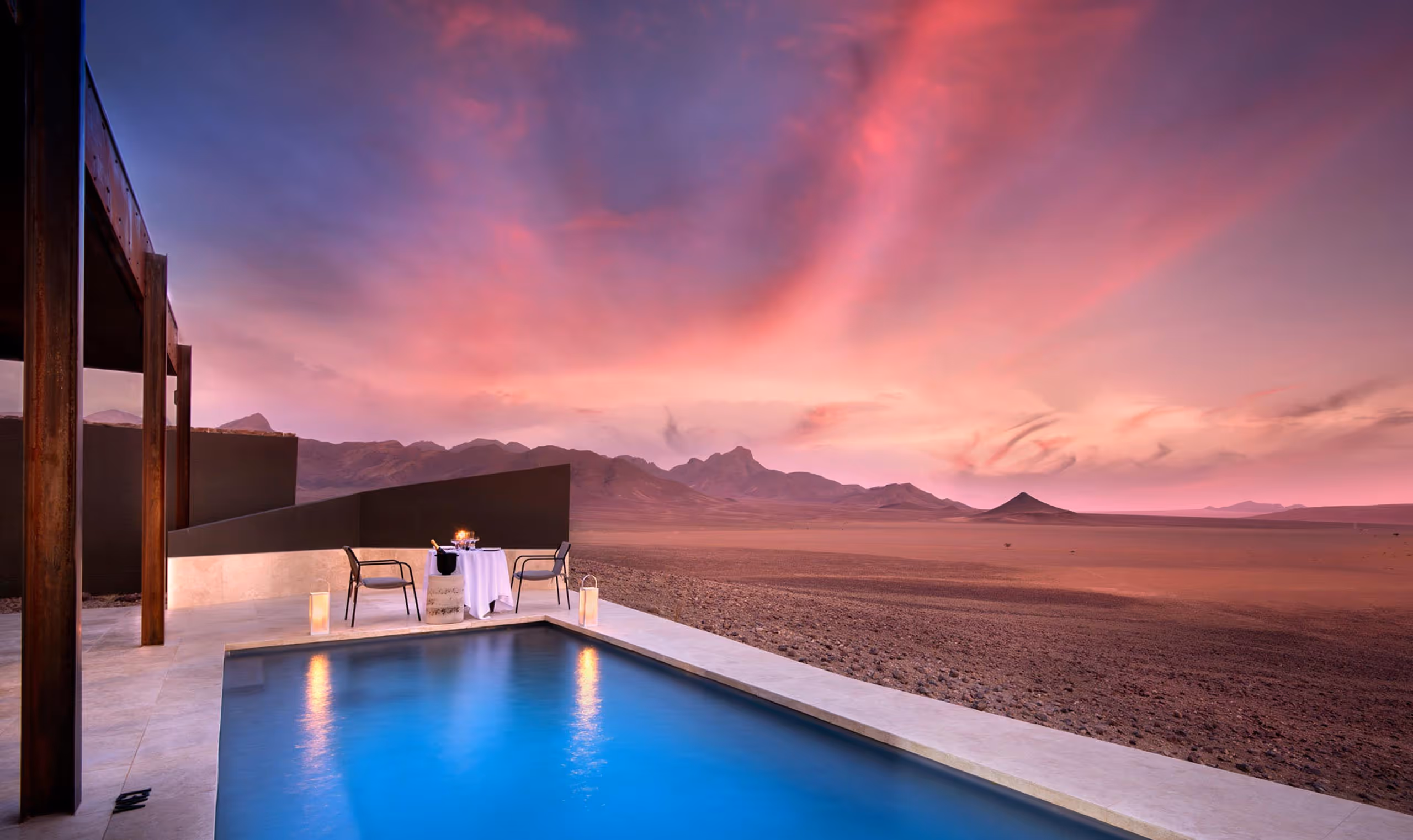 Poolside dining setup with two chairs and a table overlooking vast desert and mountains at sunset.