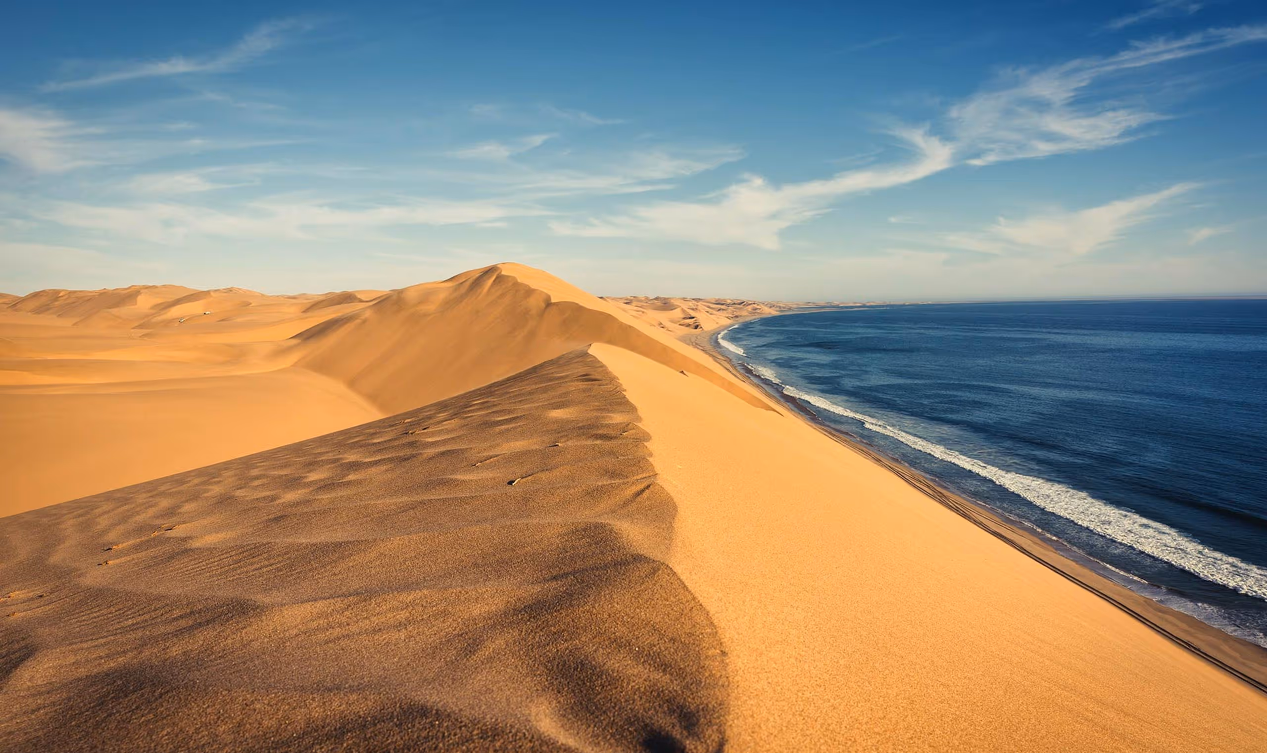 Golden sand dunes along a coastline under a partly cloudy blue sky.