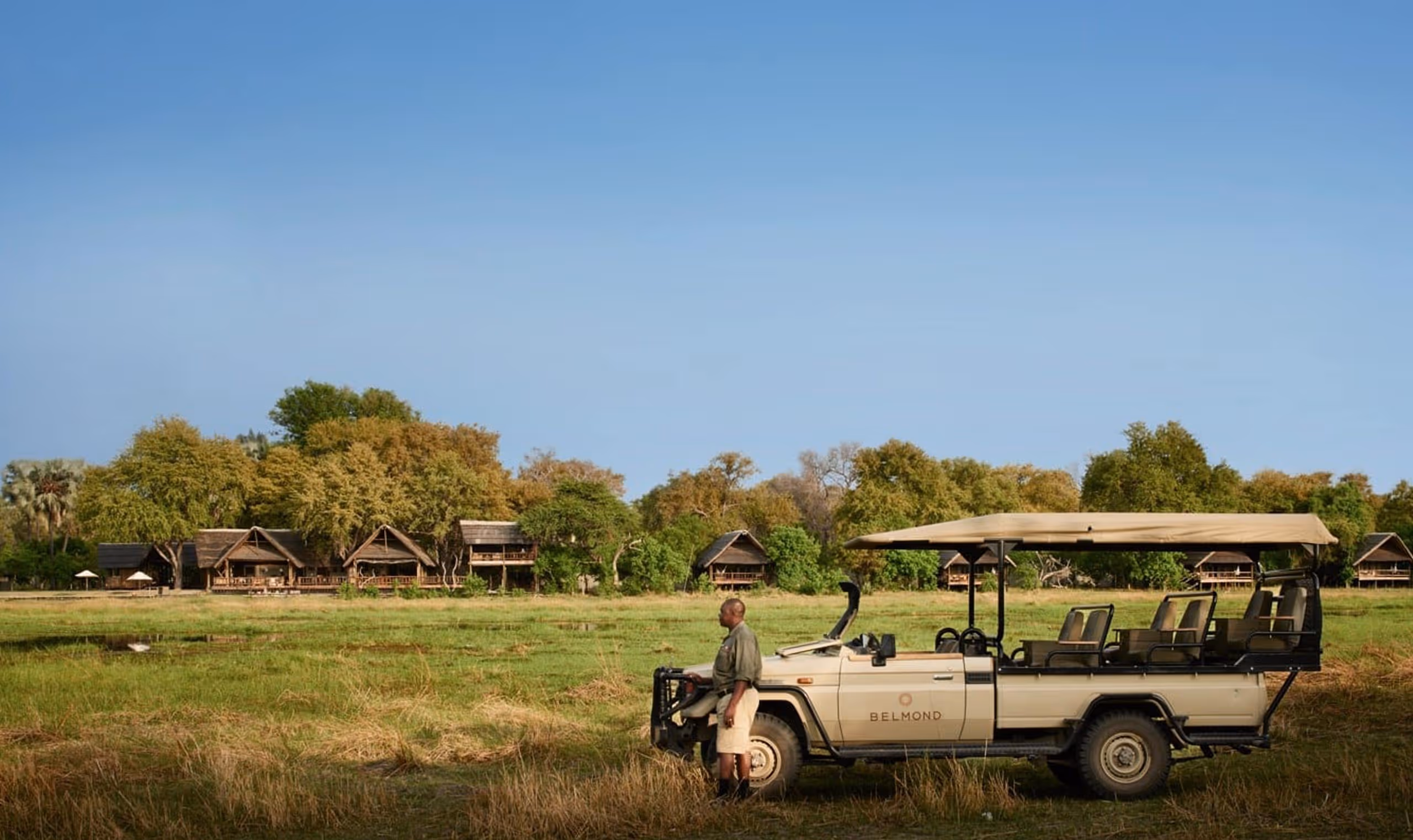 Safari guide standing by a beige open-sided safari vehicle parked in a grassy field with lodge cabins behind trees.