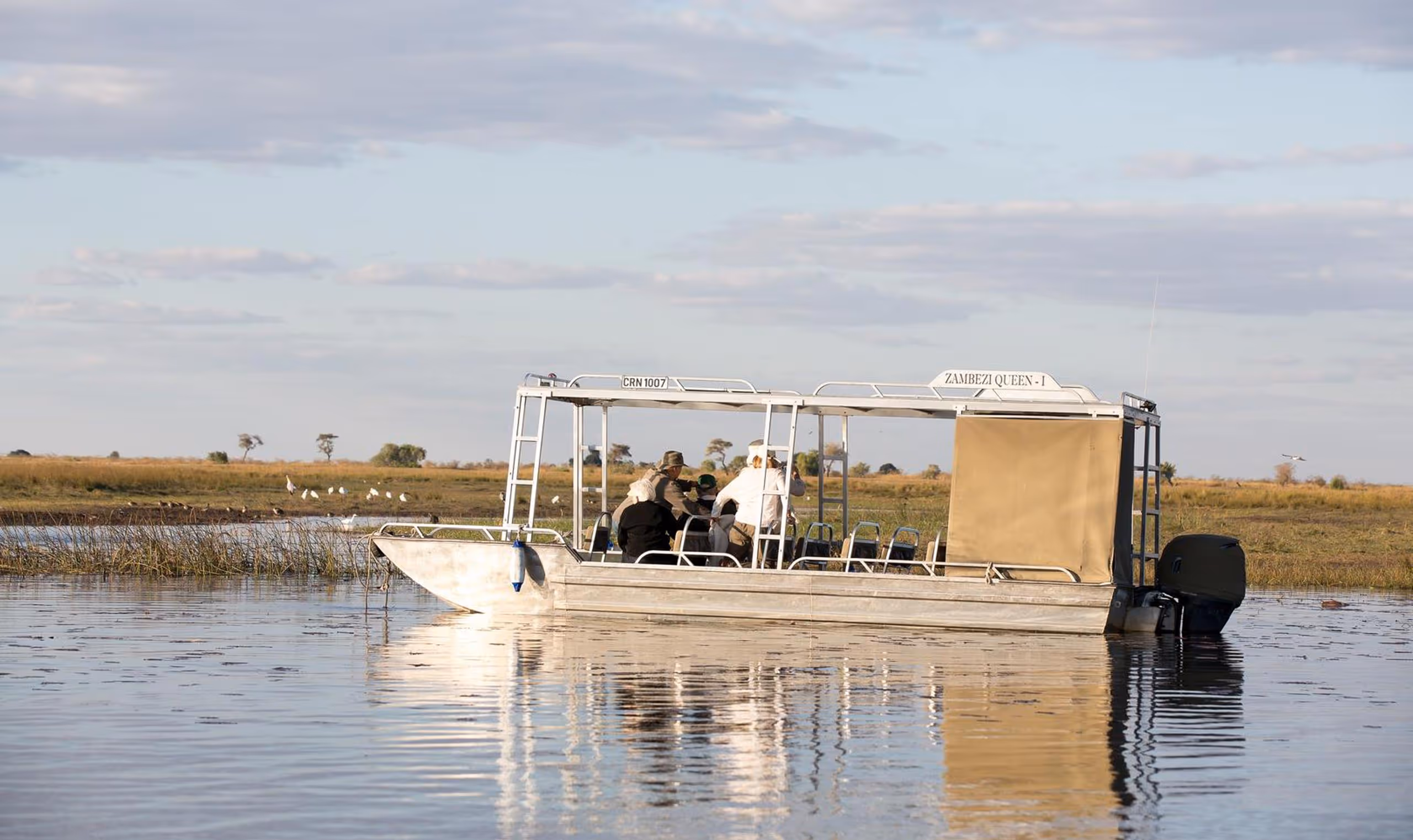 A river safari boat named Zambezi Queen-I with passengers floating on calm water near grassy banks.