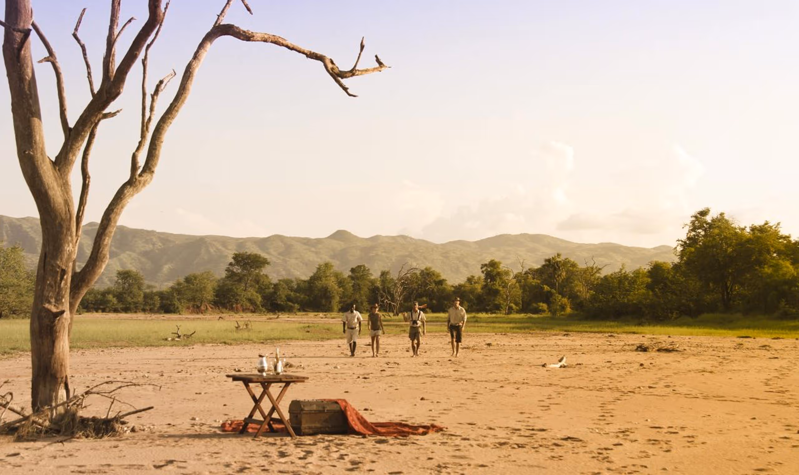 Four people walking on dry sandy land with a table and tree, green trees and hills in the distance.