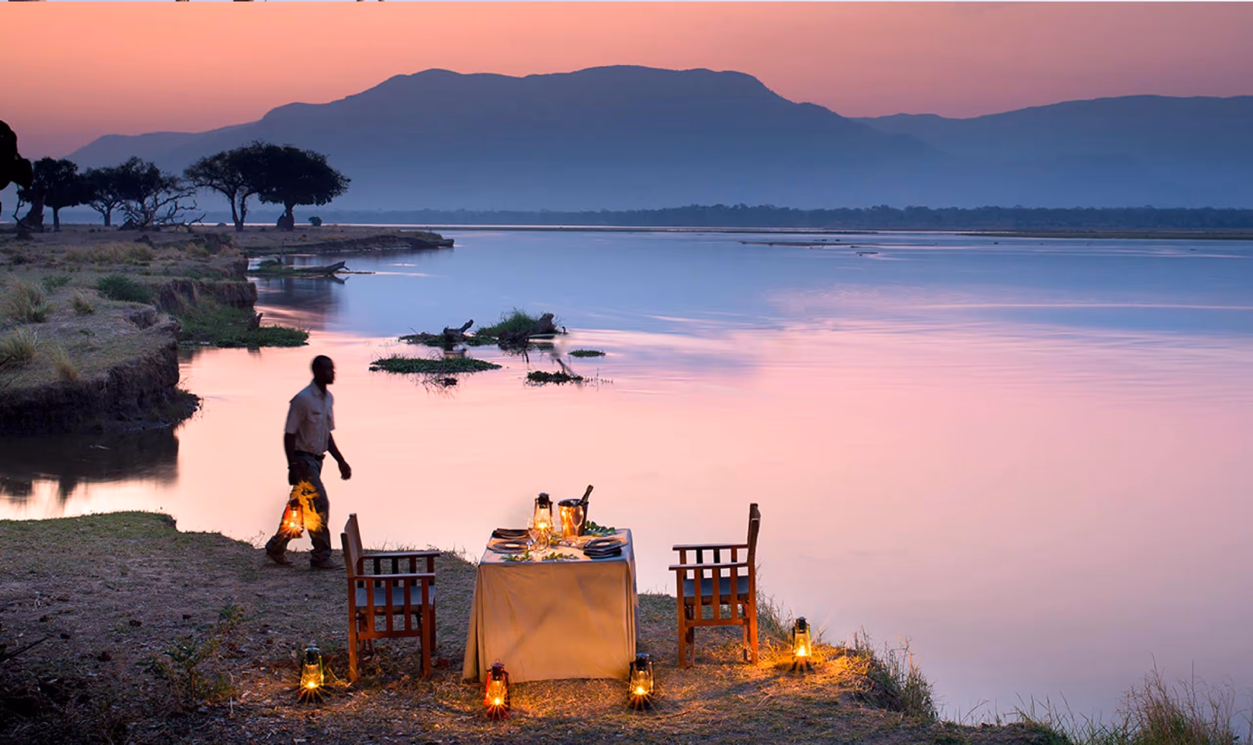 Man carrying lantern near a candlelit table set for two by a calm river at sunset with mountains in the background.