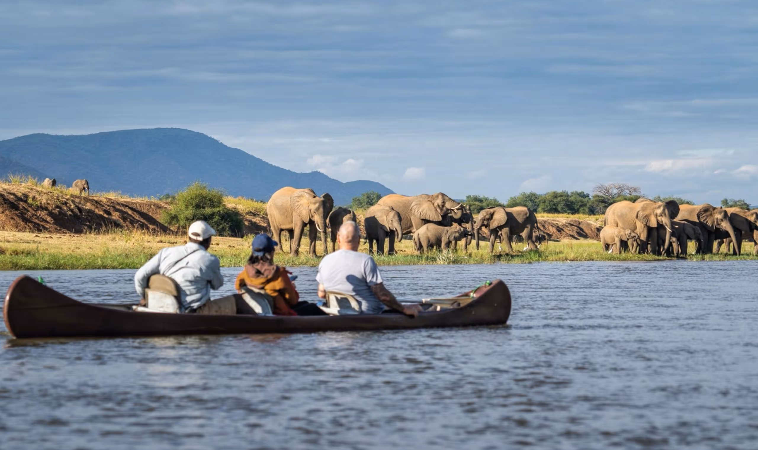 Three people in a canoe watching a herd of elephants by a riverbank with hills in the background.