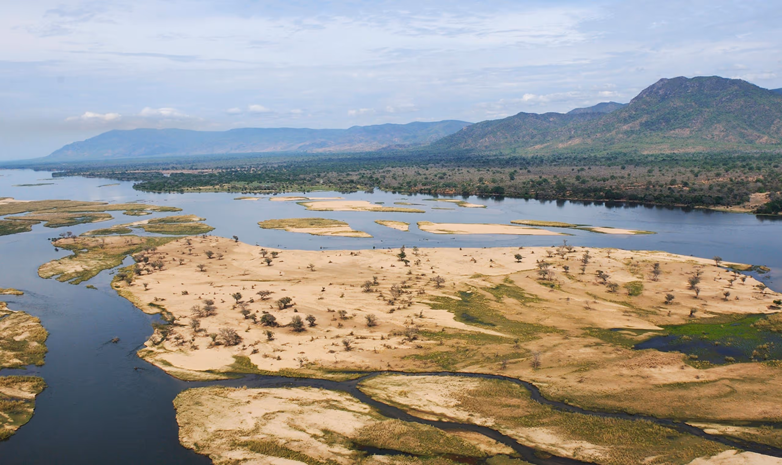 Aerial view of river islands with patches of trees and distant mountain range under a cloudy sky.