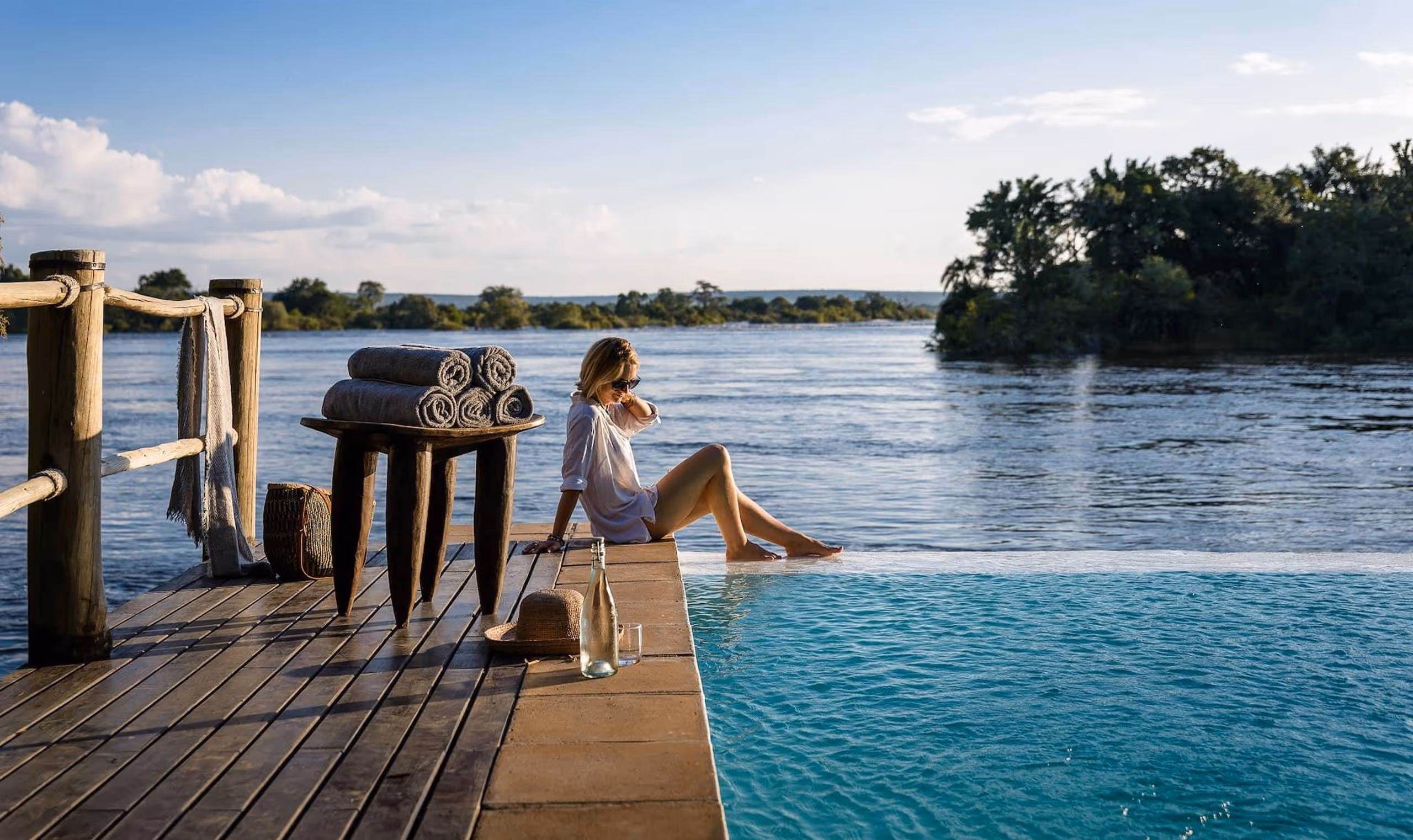 Woman sitting at edge of pool dipping feet while overlooking a river with trees and blue sky.