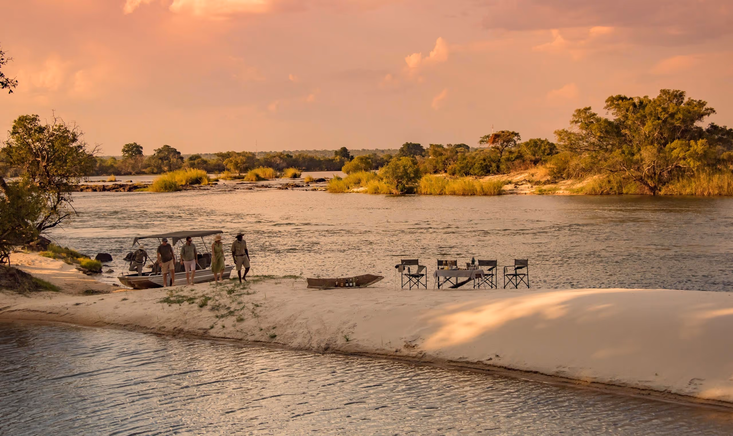 Four people walking from a boat on a sandy shore beside a river with chairs and table set up.
