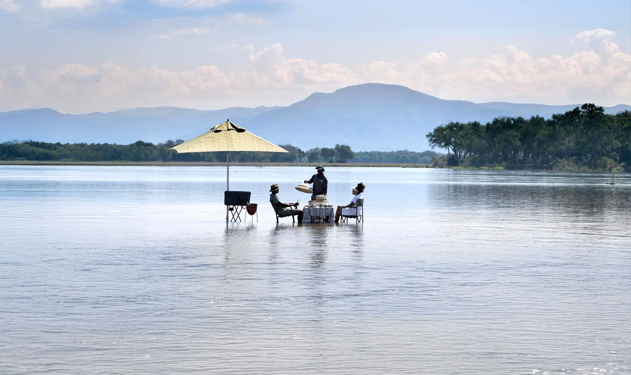 Two people seated and one serving food under umbrella while standing in shallow river water.